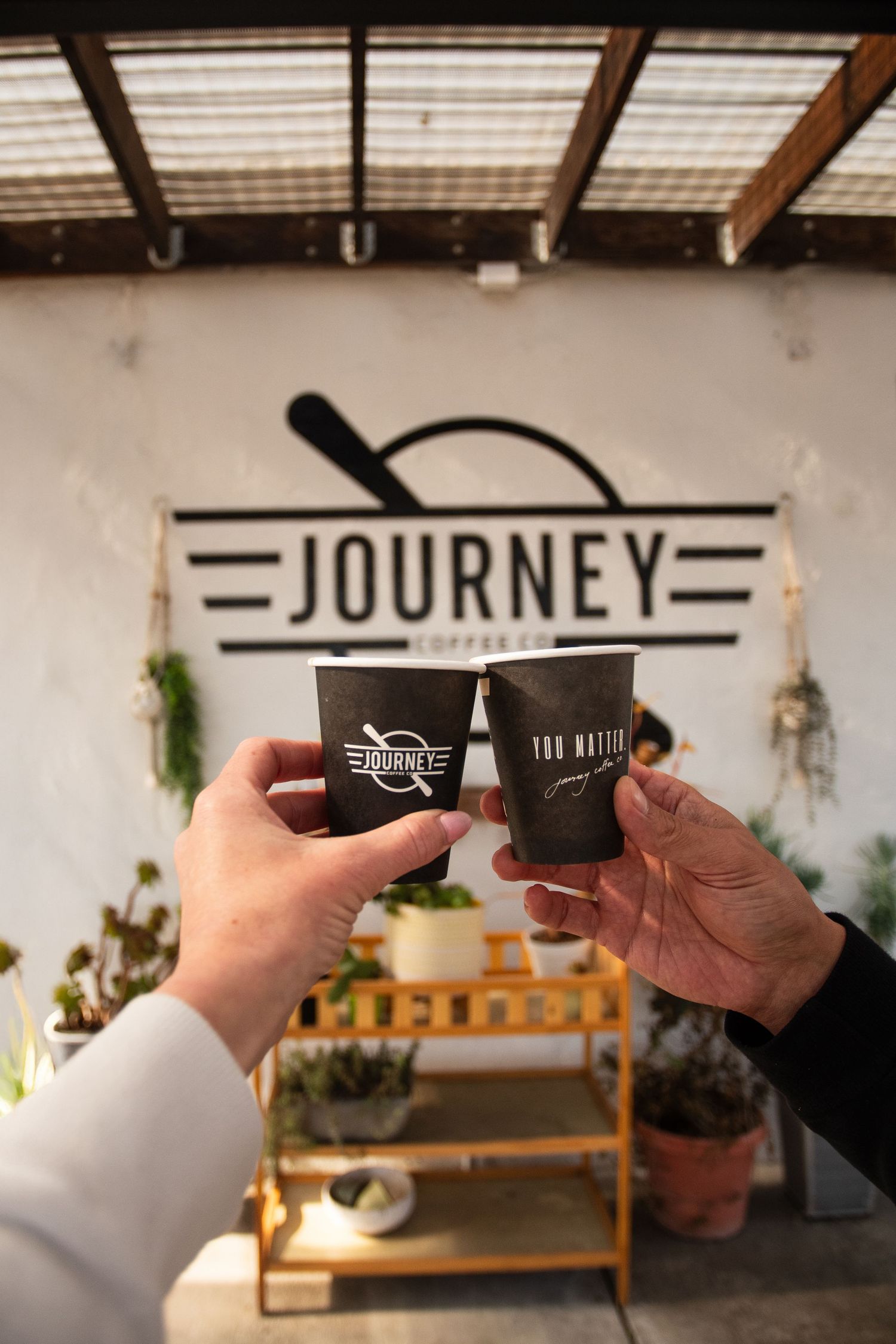 Two hands exchanging Journey Coffee Co branded cups in front of cafe signage.