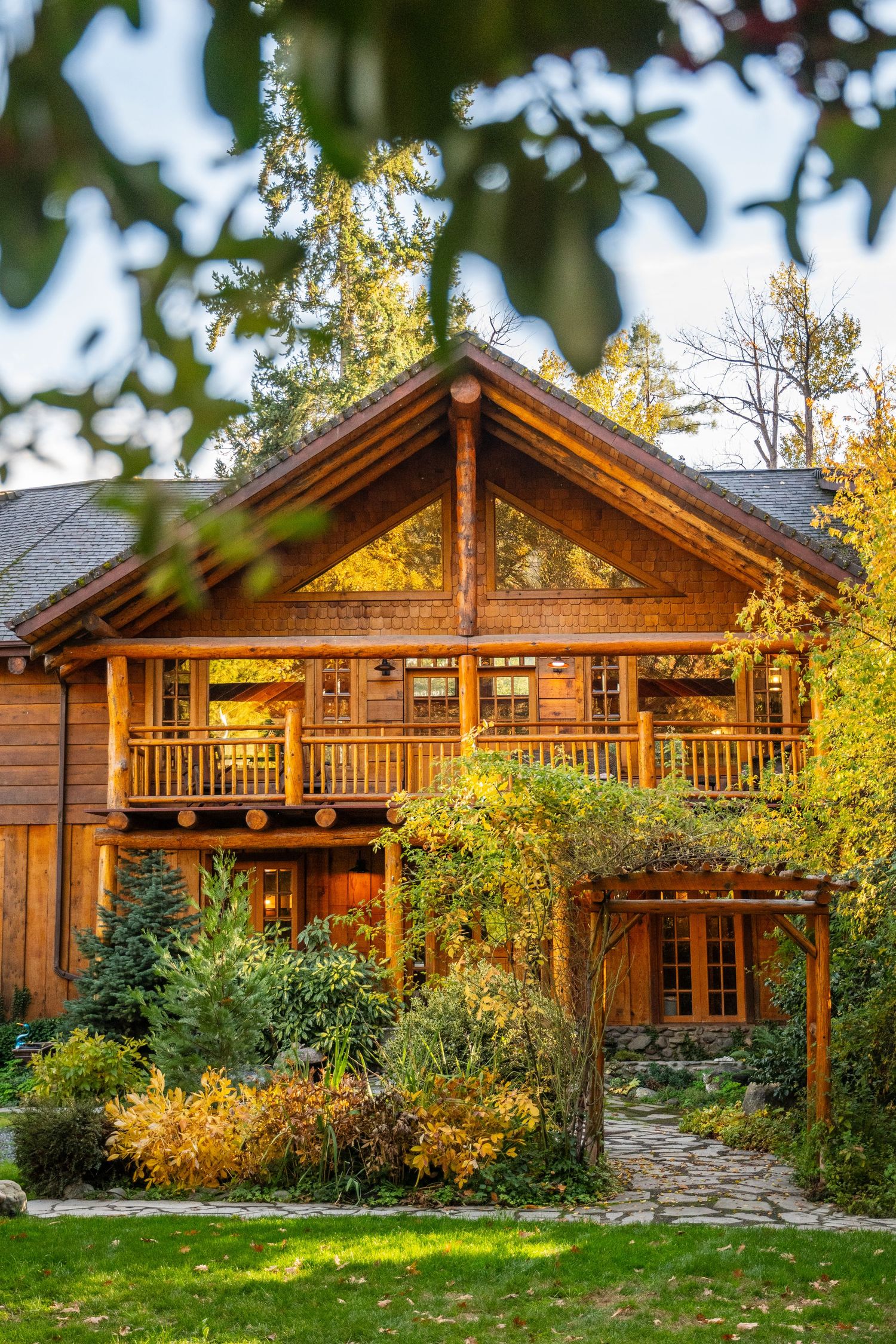 Rustic two-story log cabin with wraparound porch surrounded by autumn foliage.