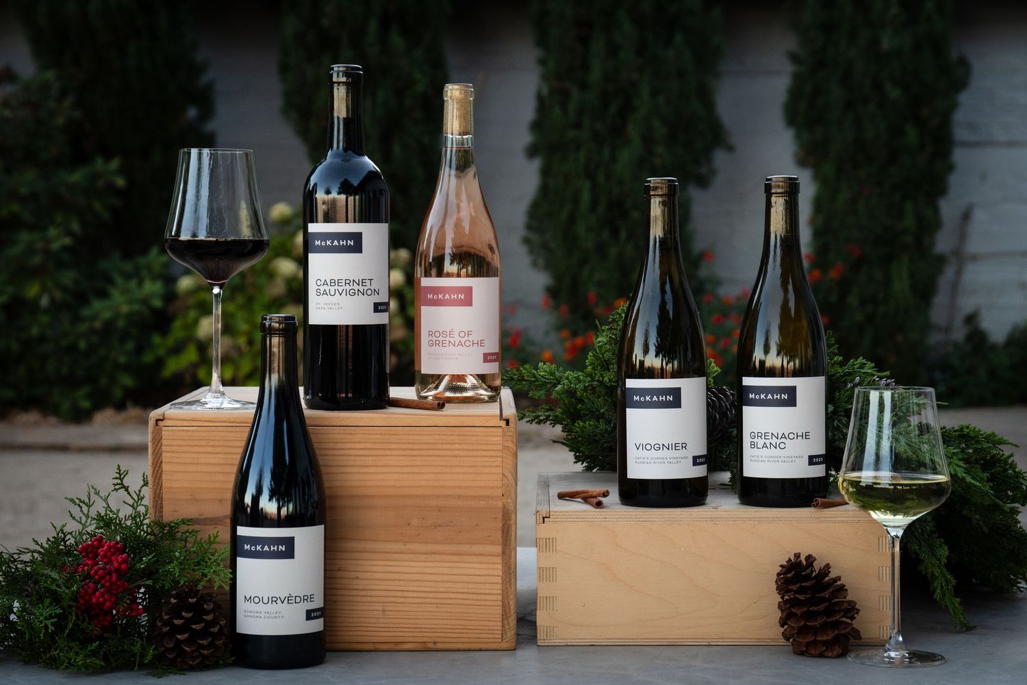 Wooden wine crates and bottles displayed outdoors against evergreen trees and white walls.
