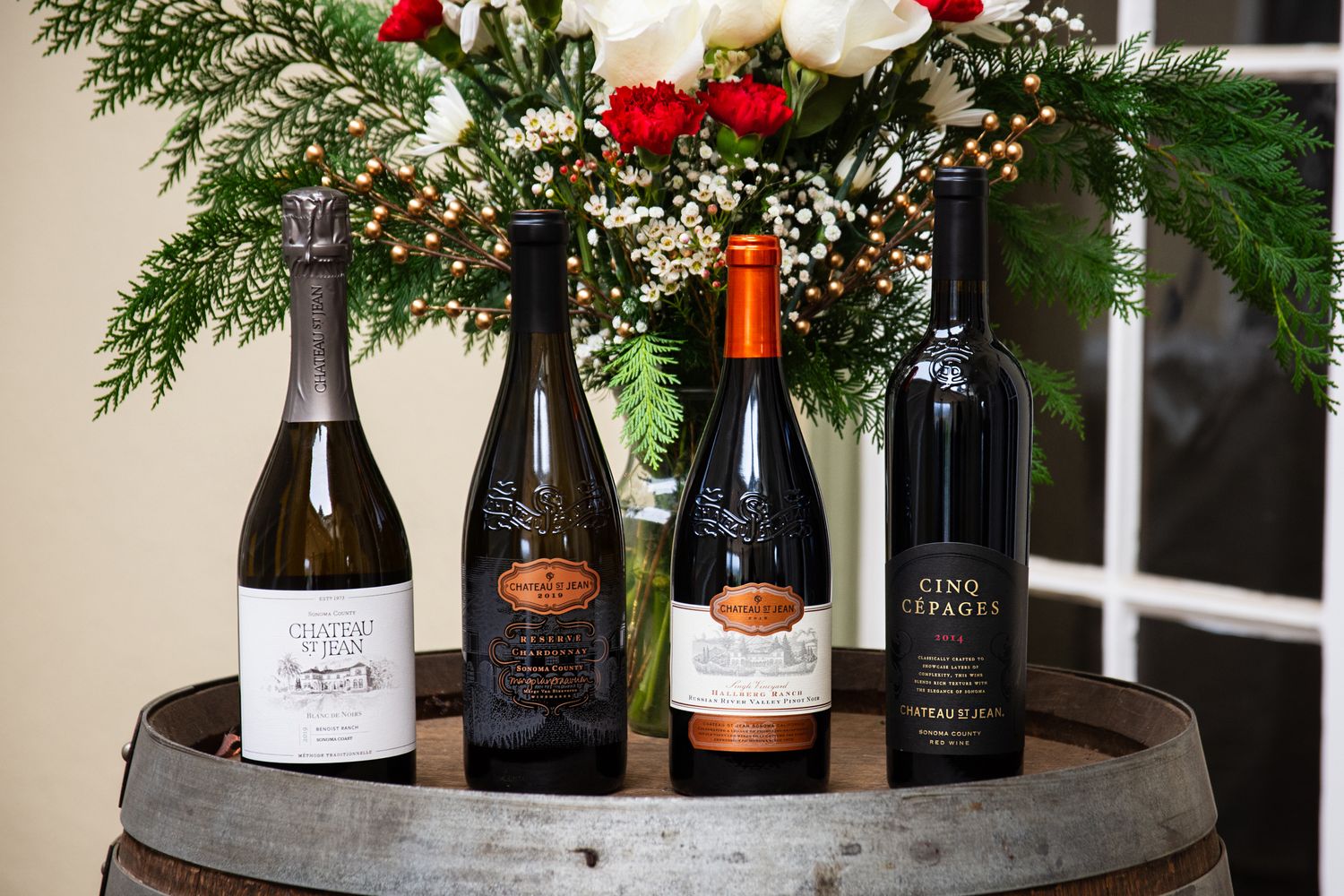 Four wine bottles arranged on wooden barrel with holiday floral arrangement.
