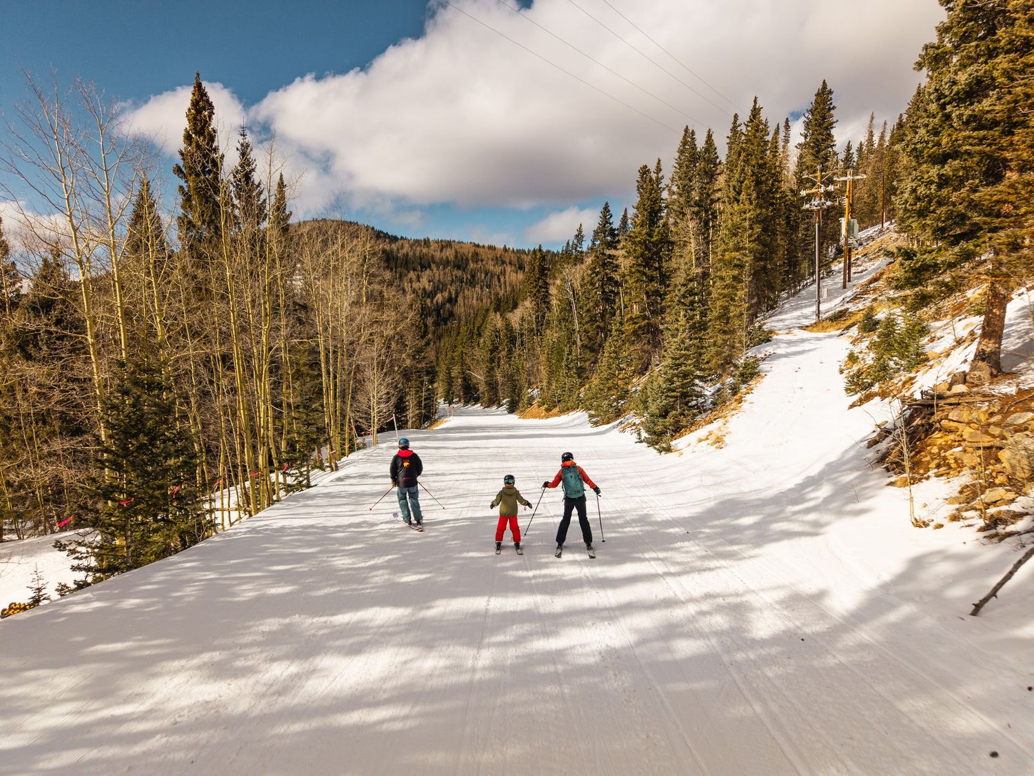 Three skiers descend a groomed ski slope surrounded by pine trees and snow-covered mountains on a sunny winter day.