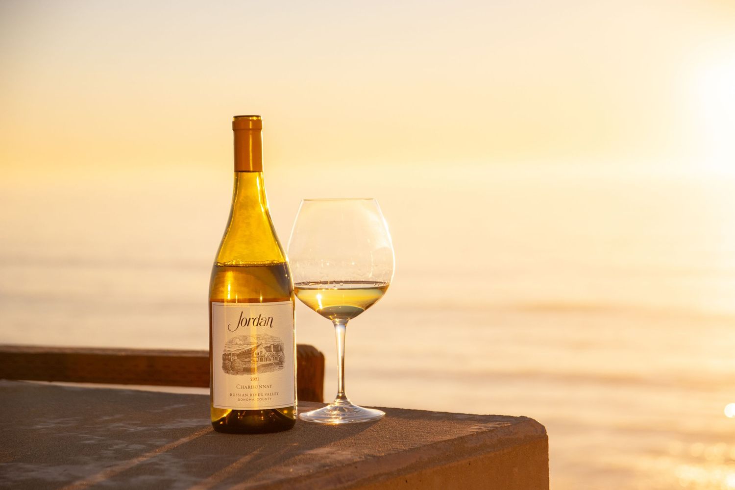 Wine glass and bottle silhouetted against a golden sunset over the ocean.