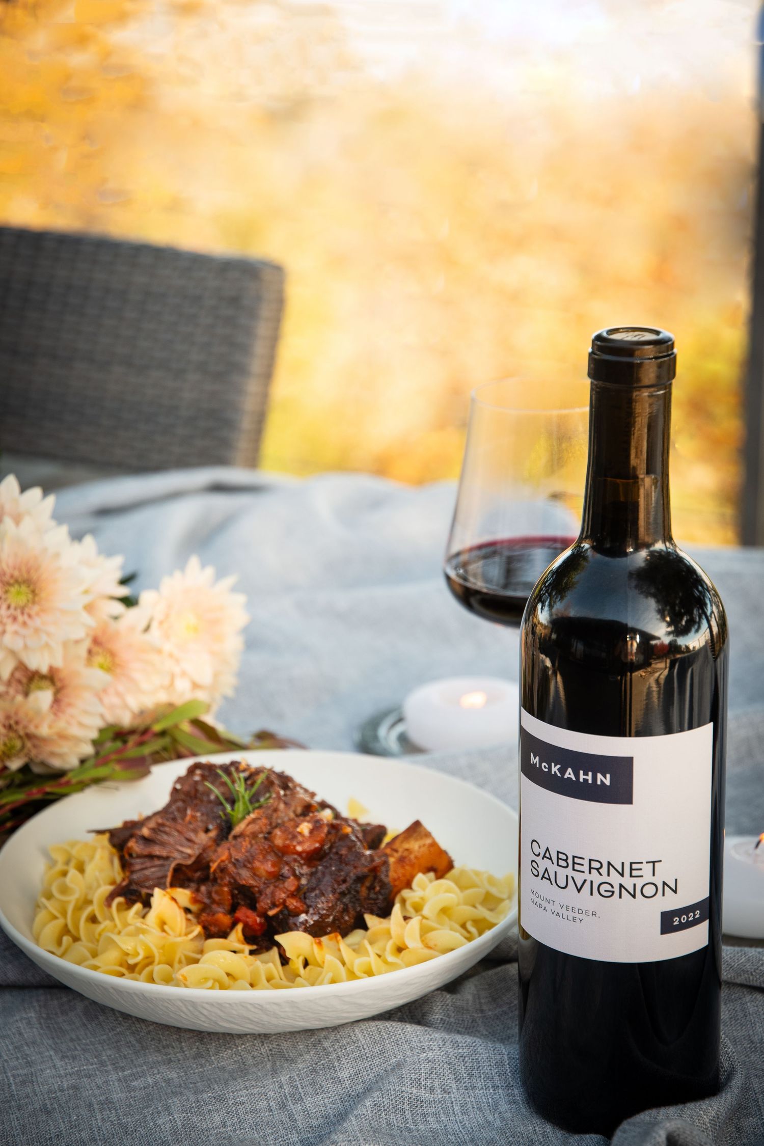 A plate of pasta with braised meat served alongside a bottle of Cabernet Sauvignon wine on an outdoor table.