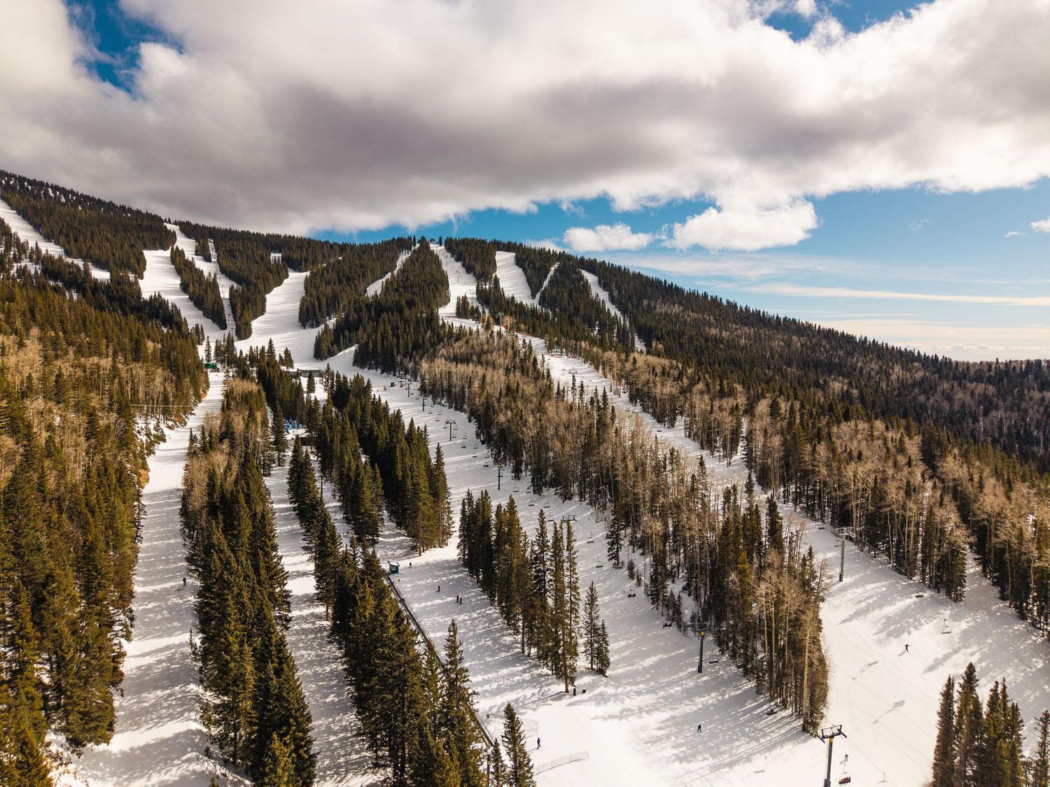 Aerial view of a snow-covered ski resort with groomed runs cutting through dense evergreen forests under a partly cloudy sky.