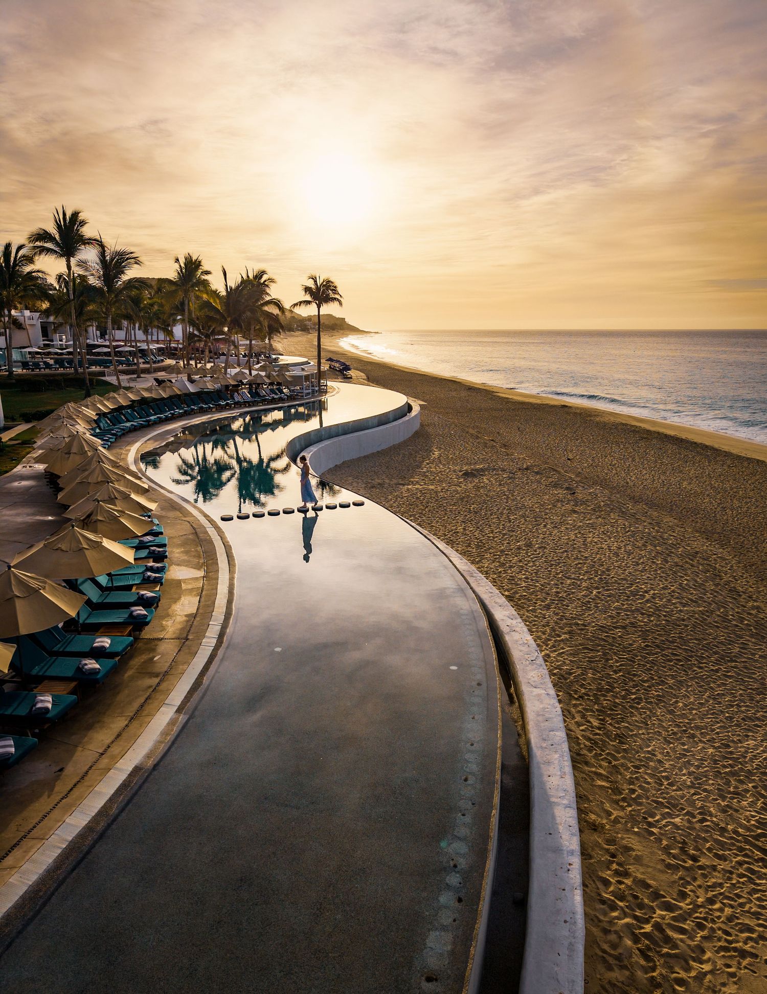 Aerial view of a luxury resort infinity pool beside a sandy beach at golden sunset with palm trees and lounge chairs.