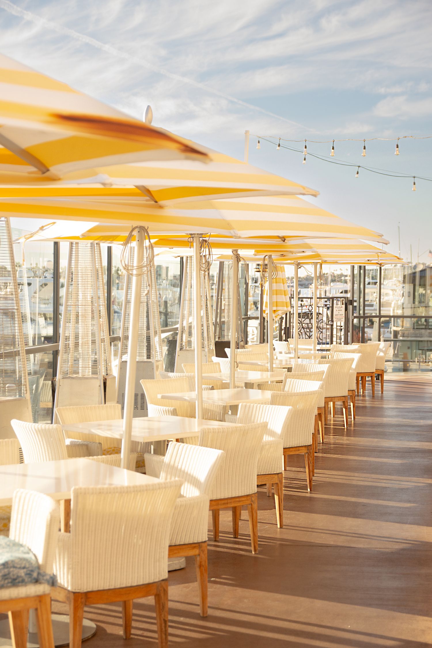 Outdoor restaurant terrace with white furniture and yellow umbrellas overlooking marina views.