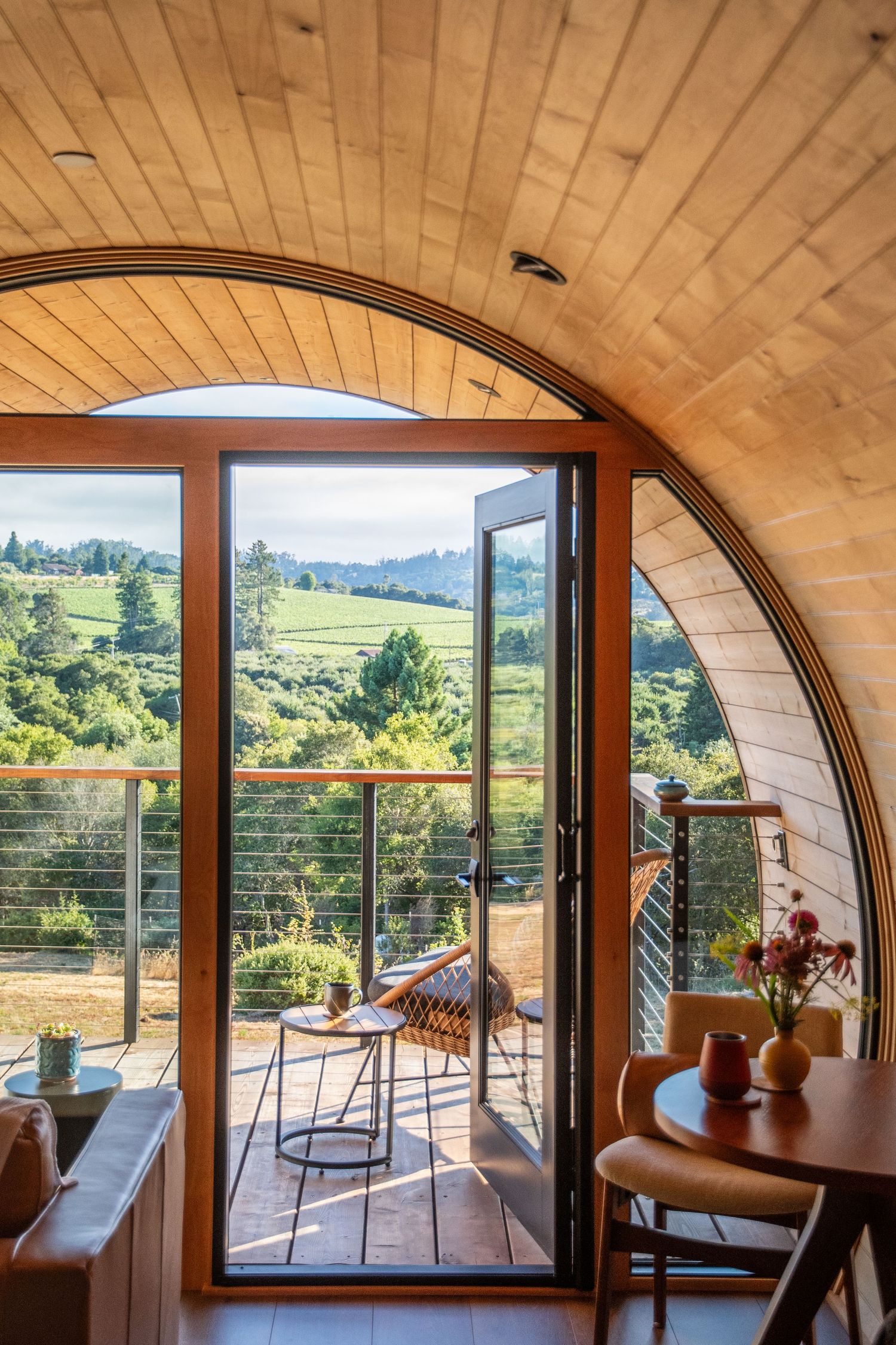 Interior of curved wooden pod with glass doors opening to a deck overlooking rolling hills.