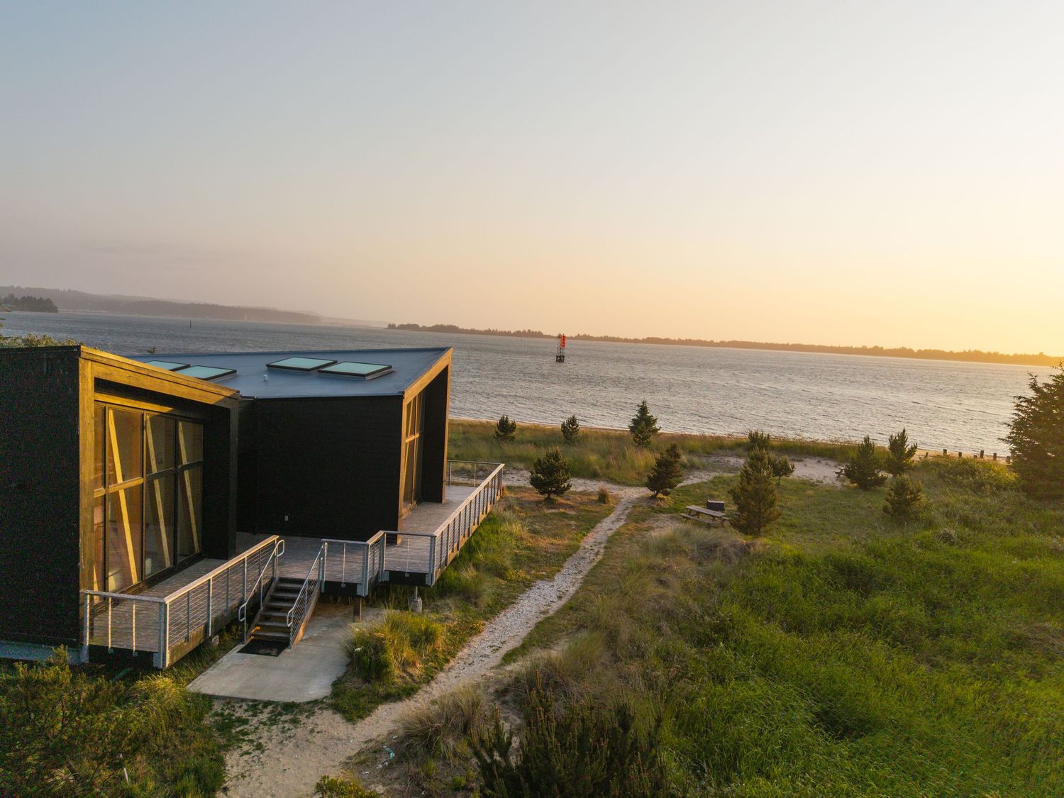 Modern coastal vacation homes with glass walls and wooden decks overlooking a scenic beach at sunset.