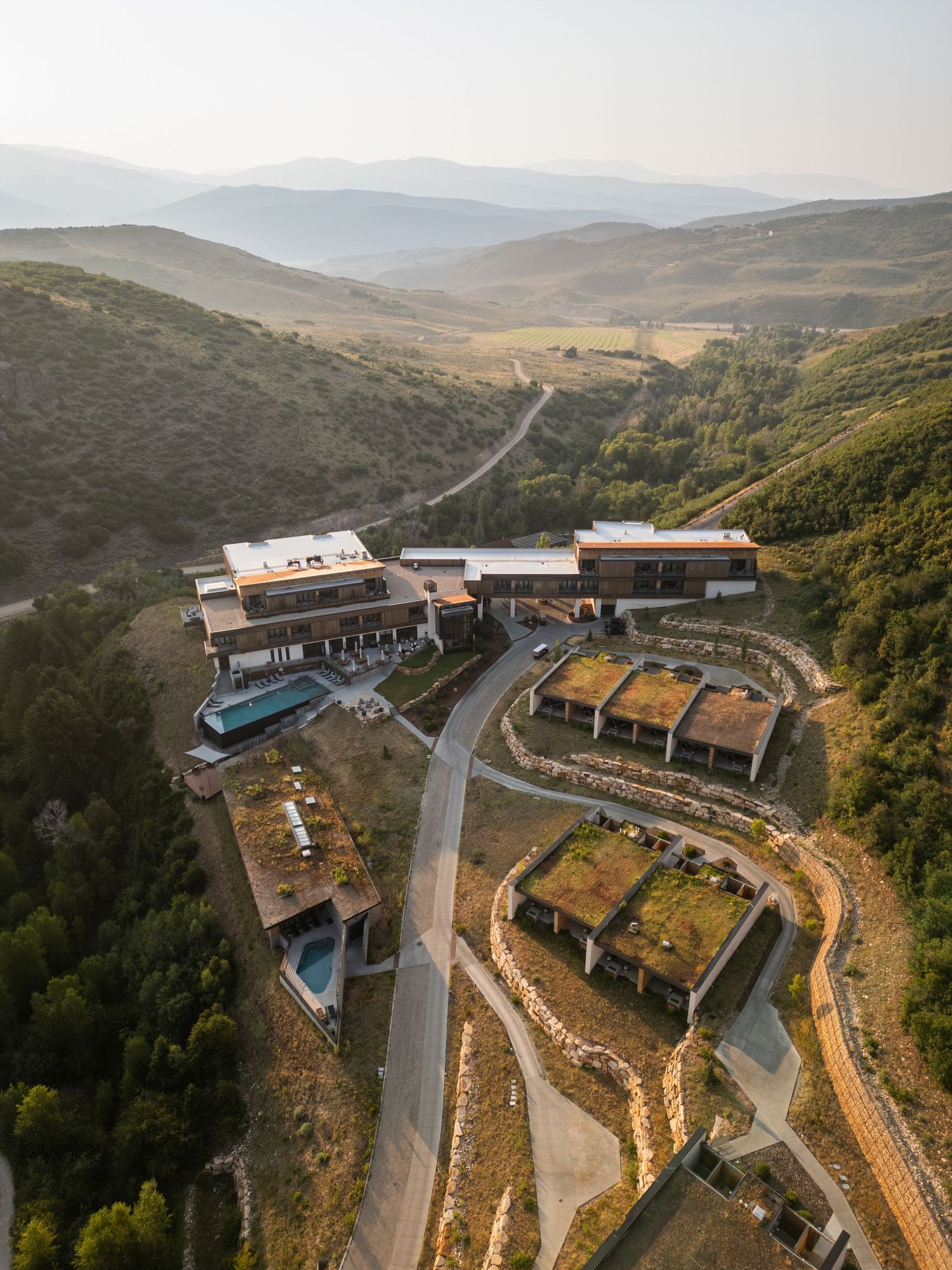 Aerial view of a modern hillside resort with green rooftops, pool, and winding roads amid lush mountains.