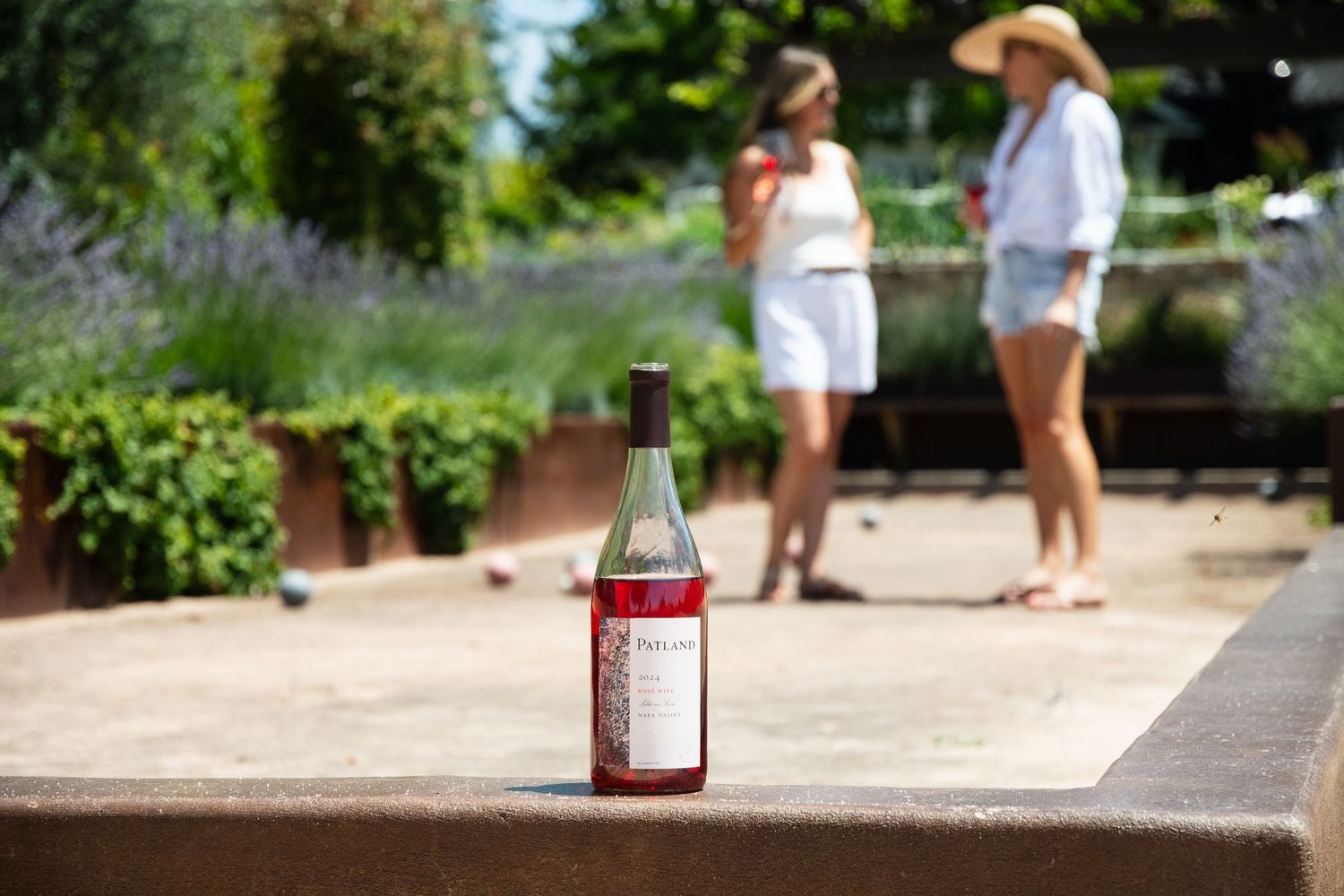 Rose wine bottle in foreground with two women enjoying drinks in a sunny garden setting.