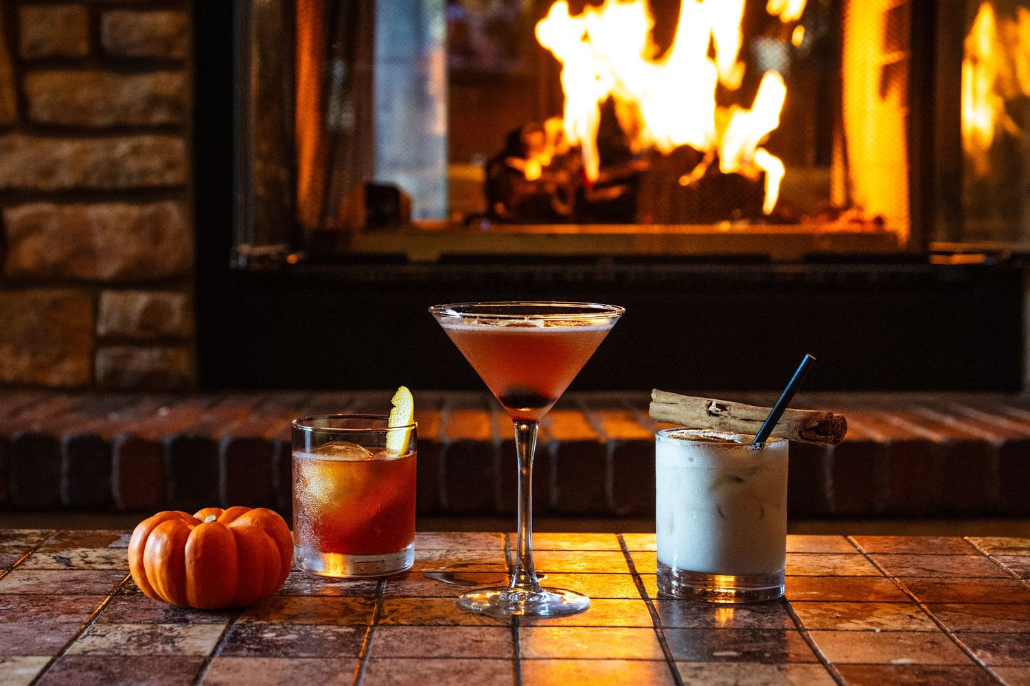 Craft cocktails and a decorative pumpkin arranged on a wooden table in front of a cozy fireplace.