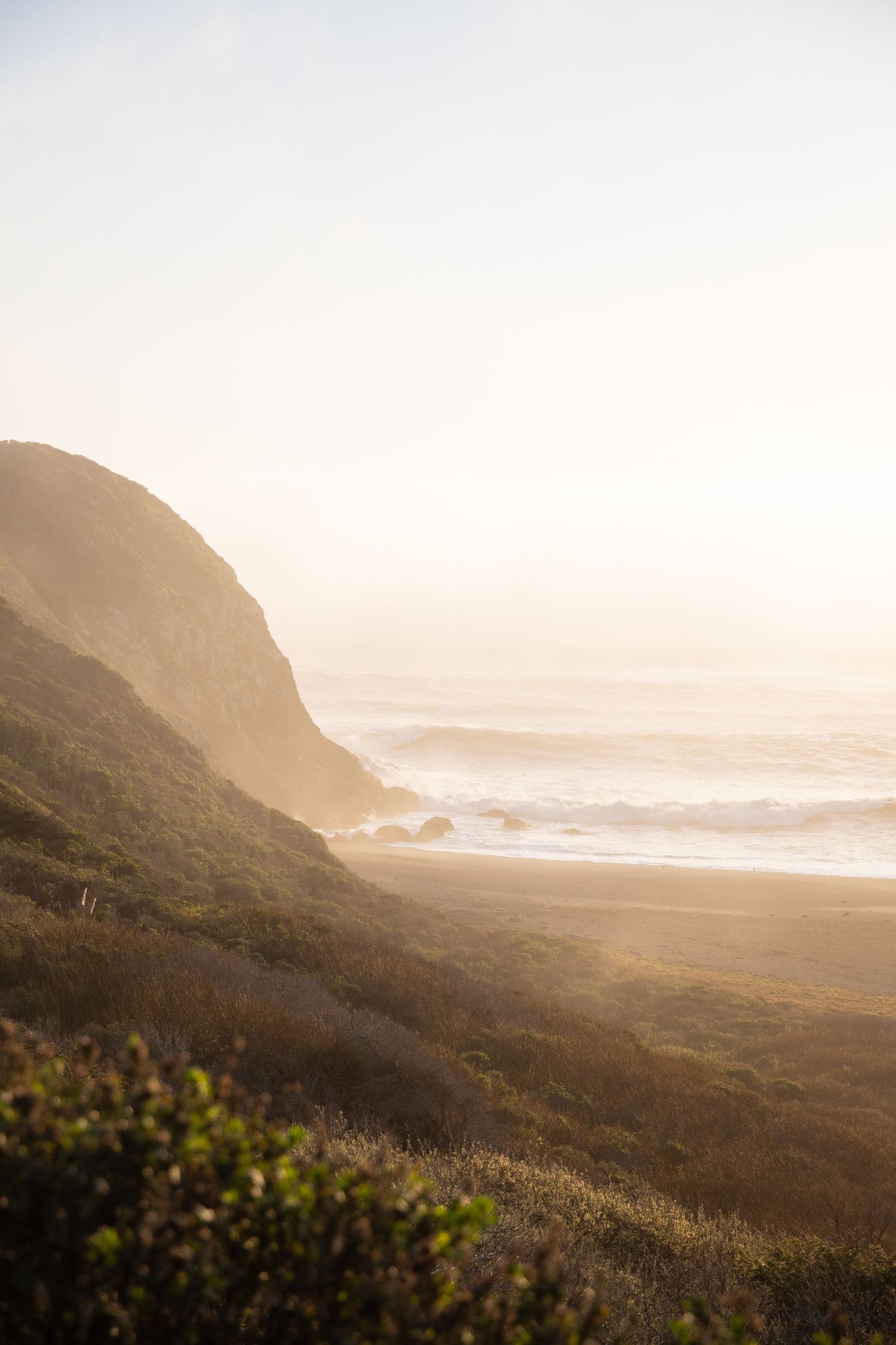 Misty coastal landscape at sunset with rolling hills and ocean waves in the distance.