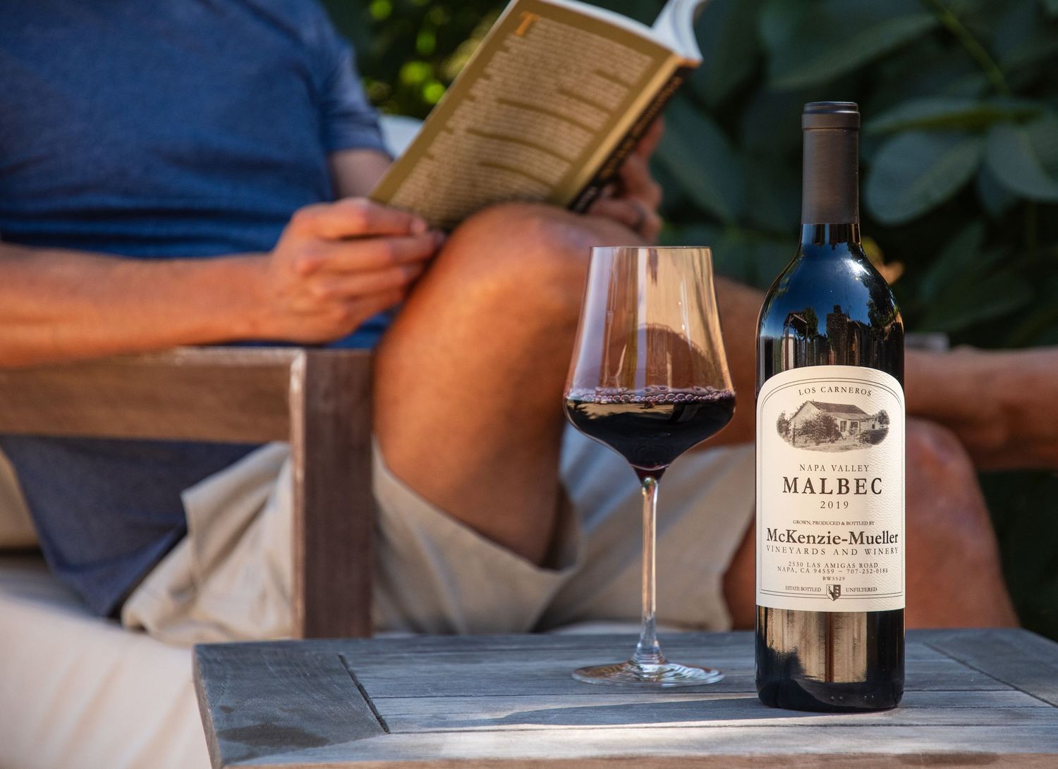 Person relaxing outdoors reading a book with a glass of Malbec red wine and bottle on a wooden table.