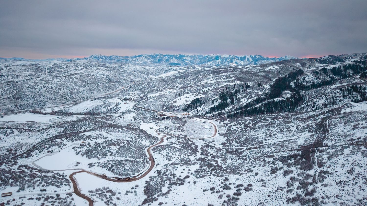 Aerial view of snow-covered mountain valley with winding road and pink-hued sky over blue peaks in winter.