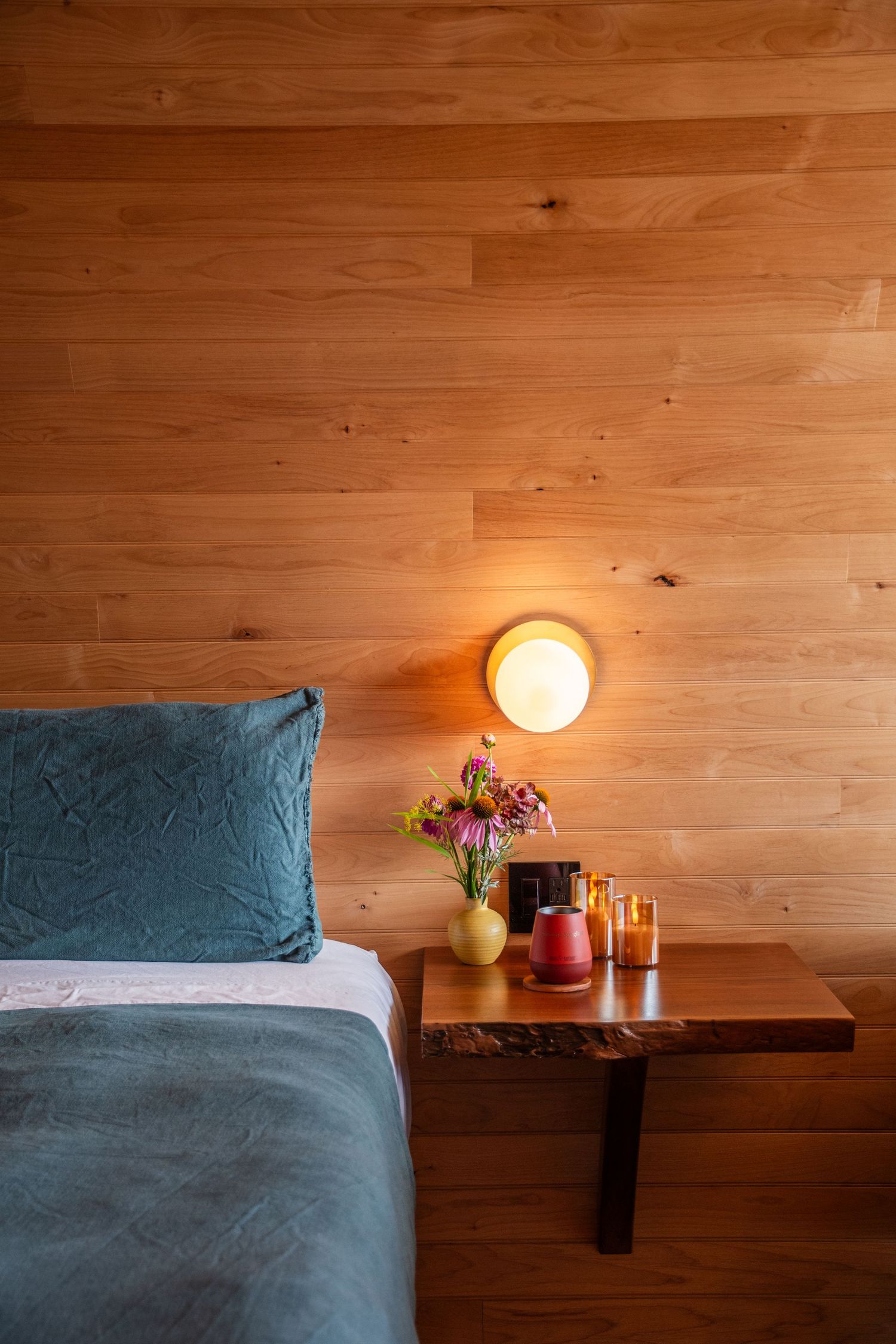 Cozy bedroom interior with wooden walls, blue bedding, and warm lighting from a round wall sconce.