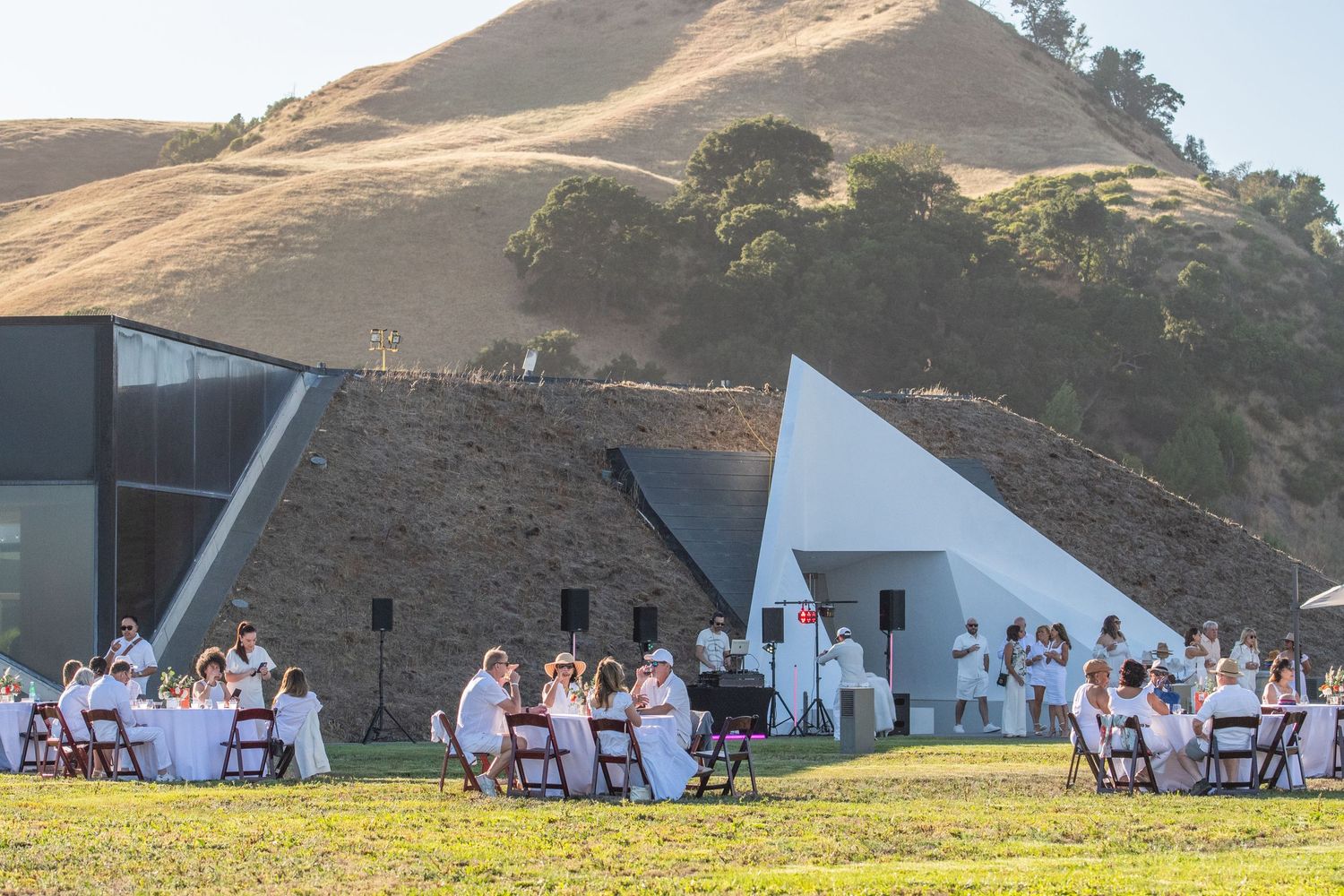 Outdoor event setup with white chairs and tables arranged on a hillside overlooking scenic mountains at sunset.