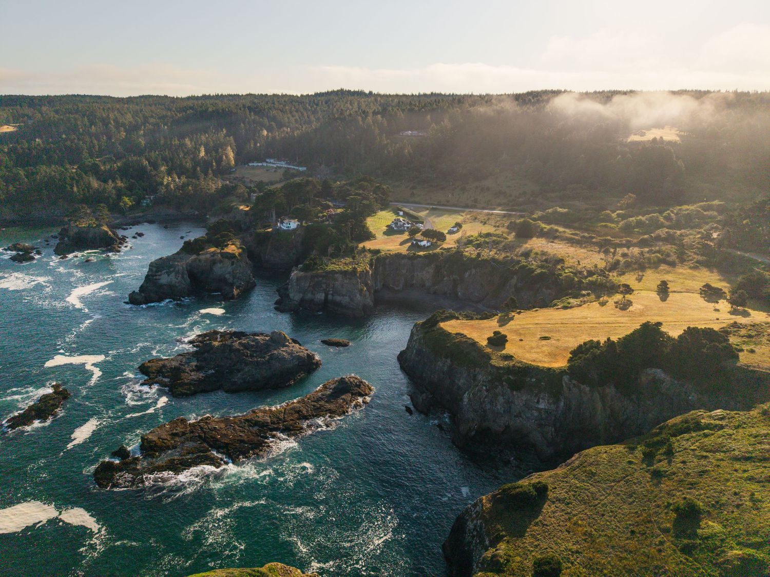 Aerial view of dramatic rocky coastline with crashing waves and green cliffs during golden hour.