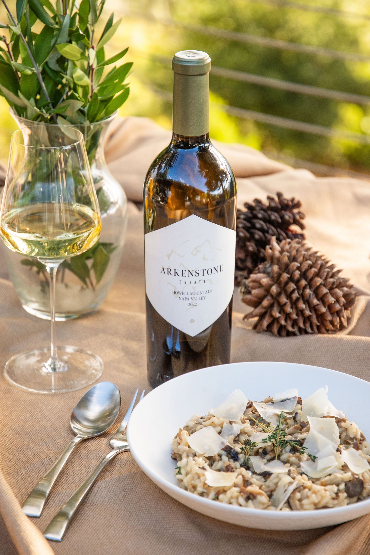 Elegant dining setup with wine bottle, glasses and risotto dish decorated with pine cones.