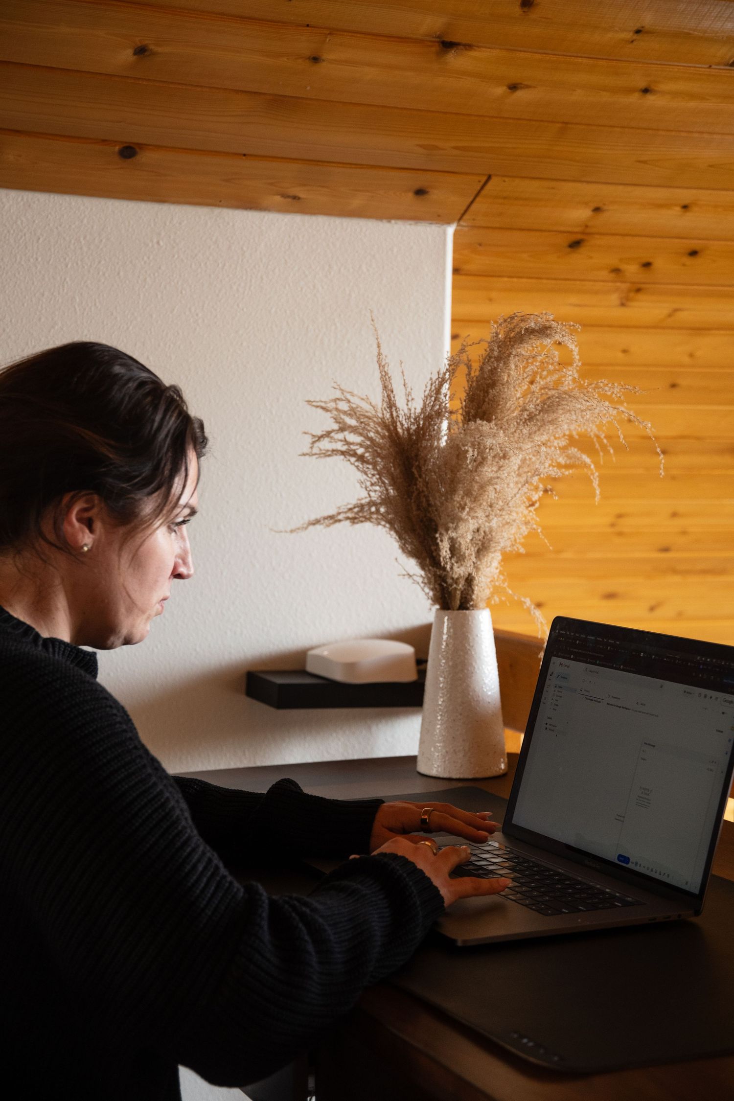 Woman in black sweater working on laptop at wooden desk with decorative dried pampas grass vase.