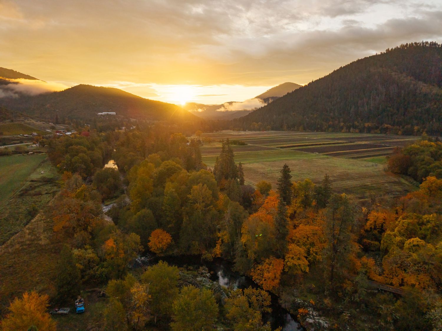 Aerial autumn sunset over a mountain valley with colorful trees, a winding river, green fields, and misty peaks.
