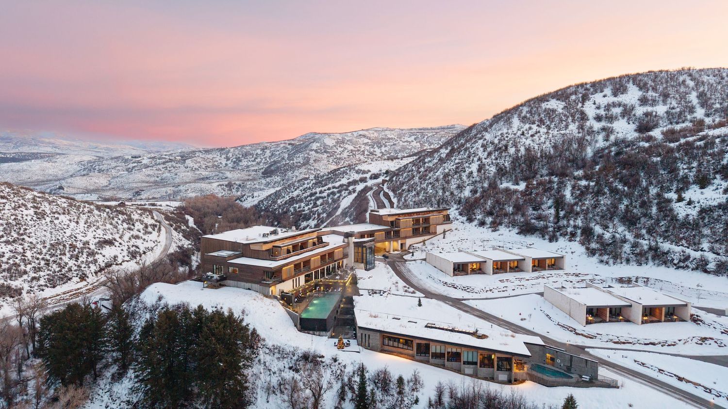 Luxury mountain resort blanketed in snow at sunset, nestled among snowy hills with a pink sky backdrop.