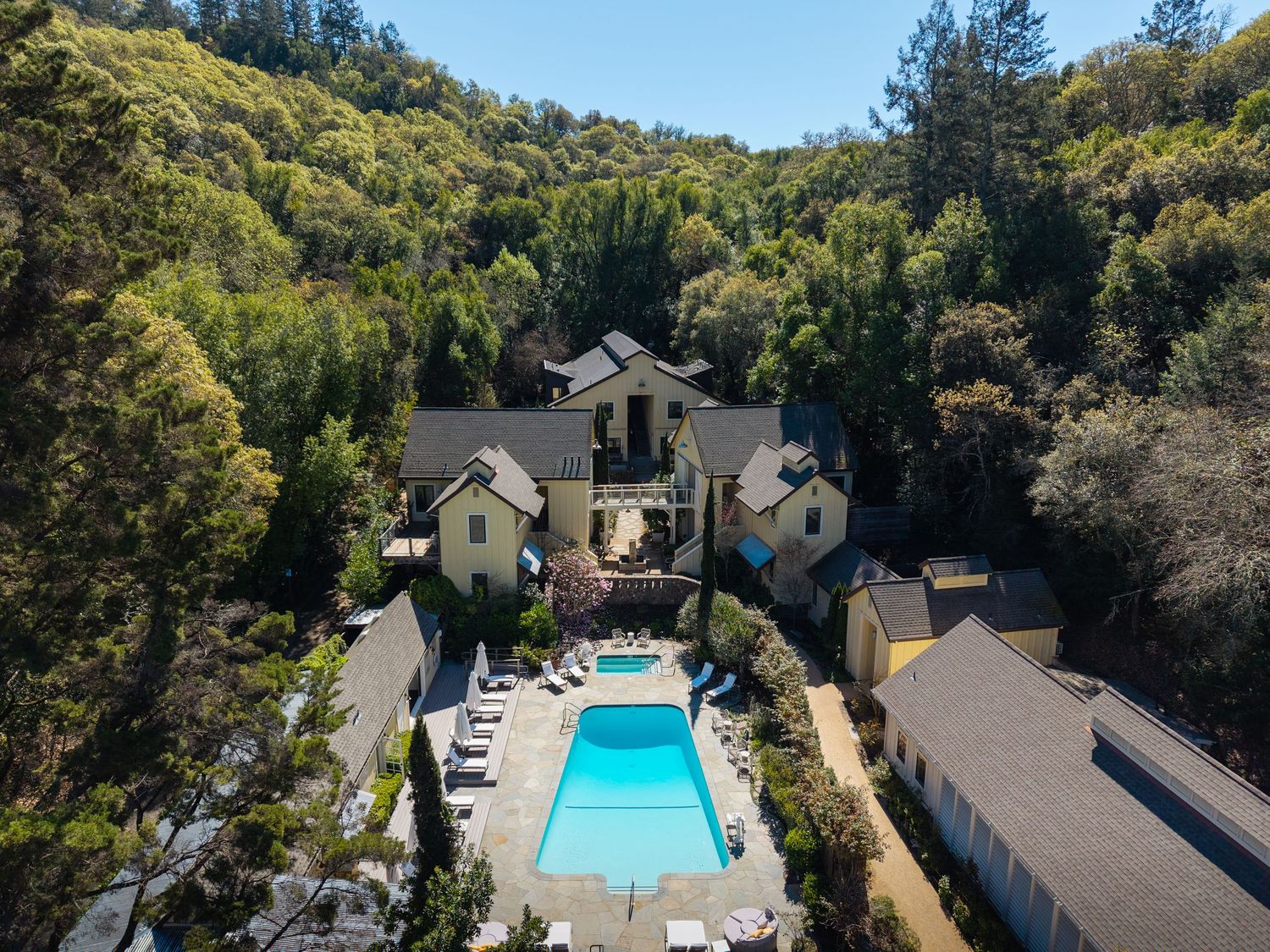 Aerial view of a luxury estate with a large swimming pool surrounded by lounge chairs, nestled among lush green hills.