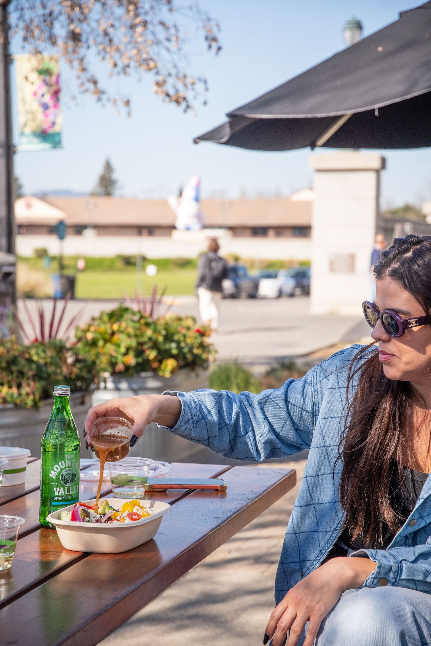 Outdoor dining scene with green glass bottle and plate of food on wooden table under umbrella.