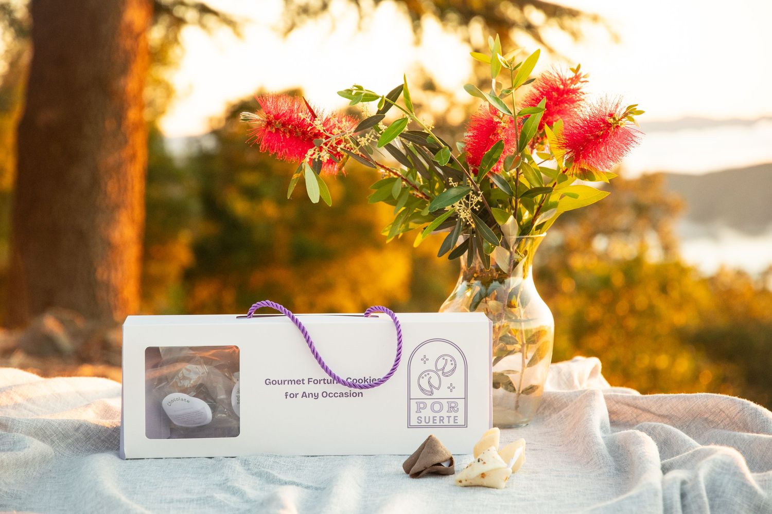 Gourmet gift box with purple ribbon beside red flowers in vase on white cloth at golden sunset outdoors.