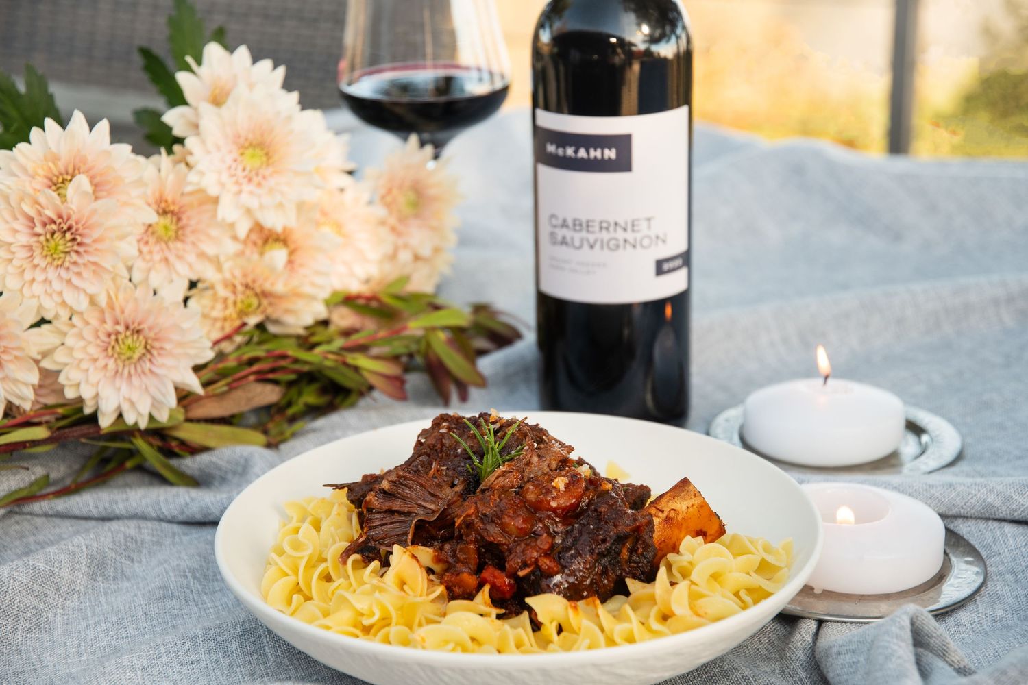 Braised meat dish served over yellow rice with red wine and pink flowers as table decoration.