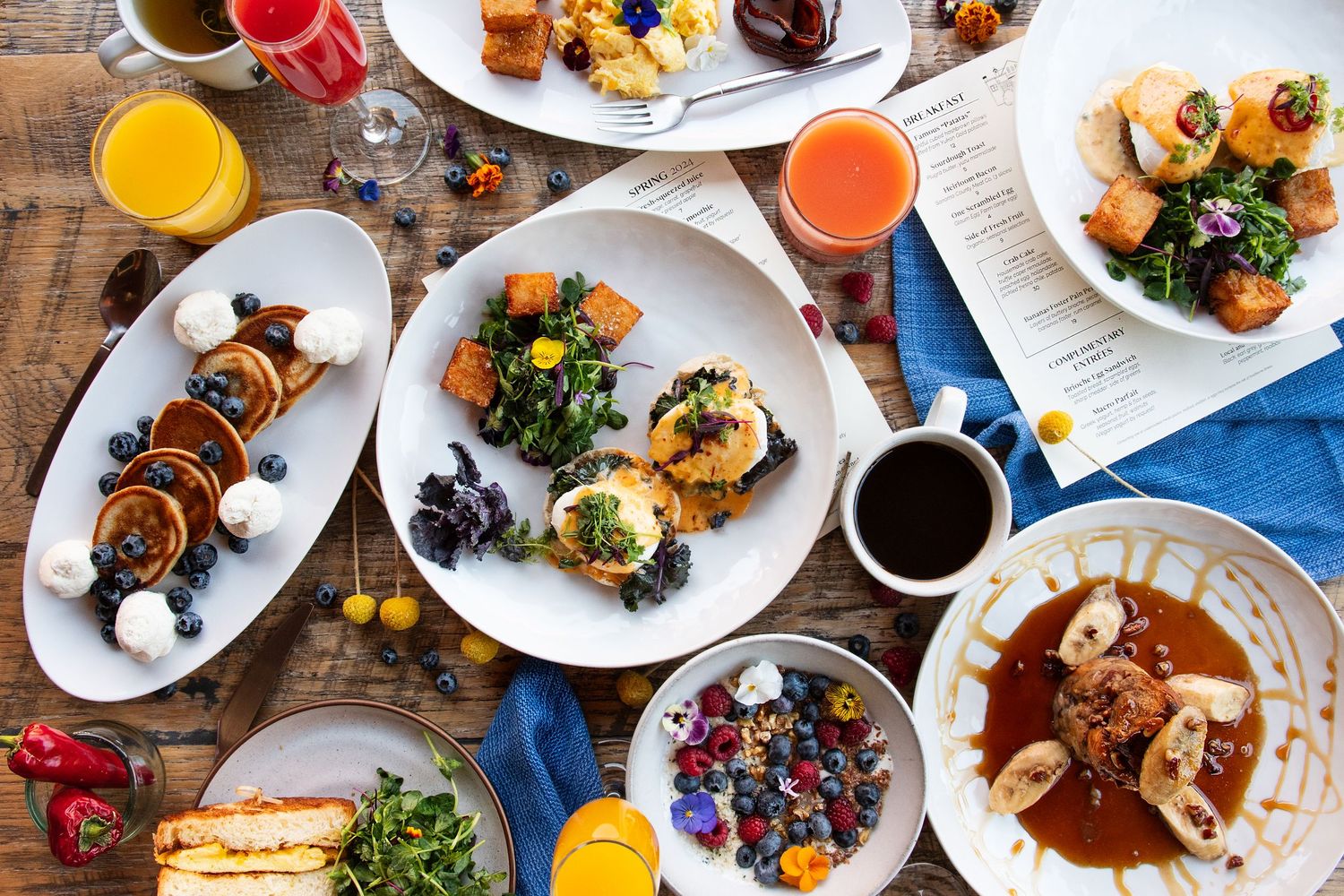 Overhead view of brunch spread with pancakes, eggs, salads and colorful drinks on wooden table.