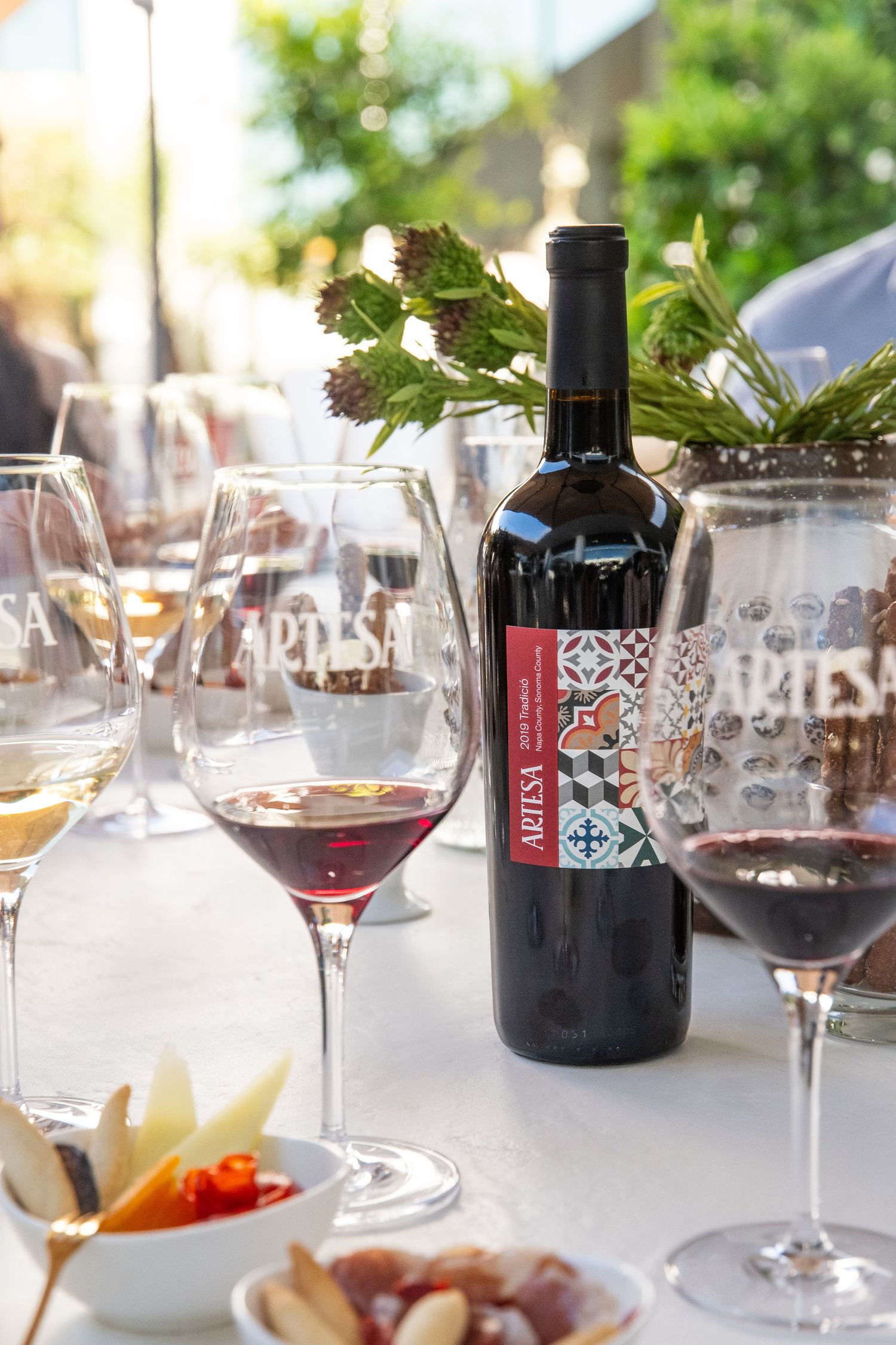 Red wine bottle and glasses on table with appetizers and grape vine decor.