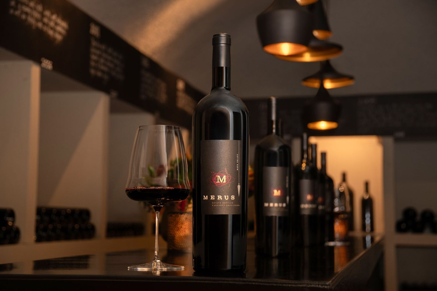 Merva wine bottles and a glass of red wine displayed on a dark table in a dimly lit cellar tasting room.