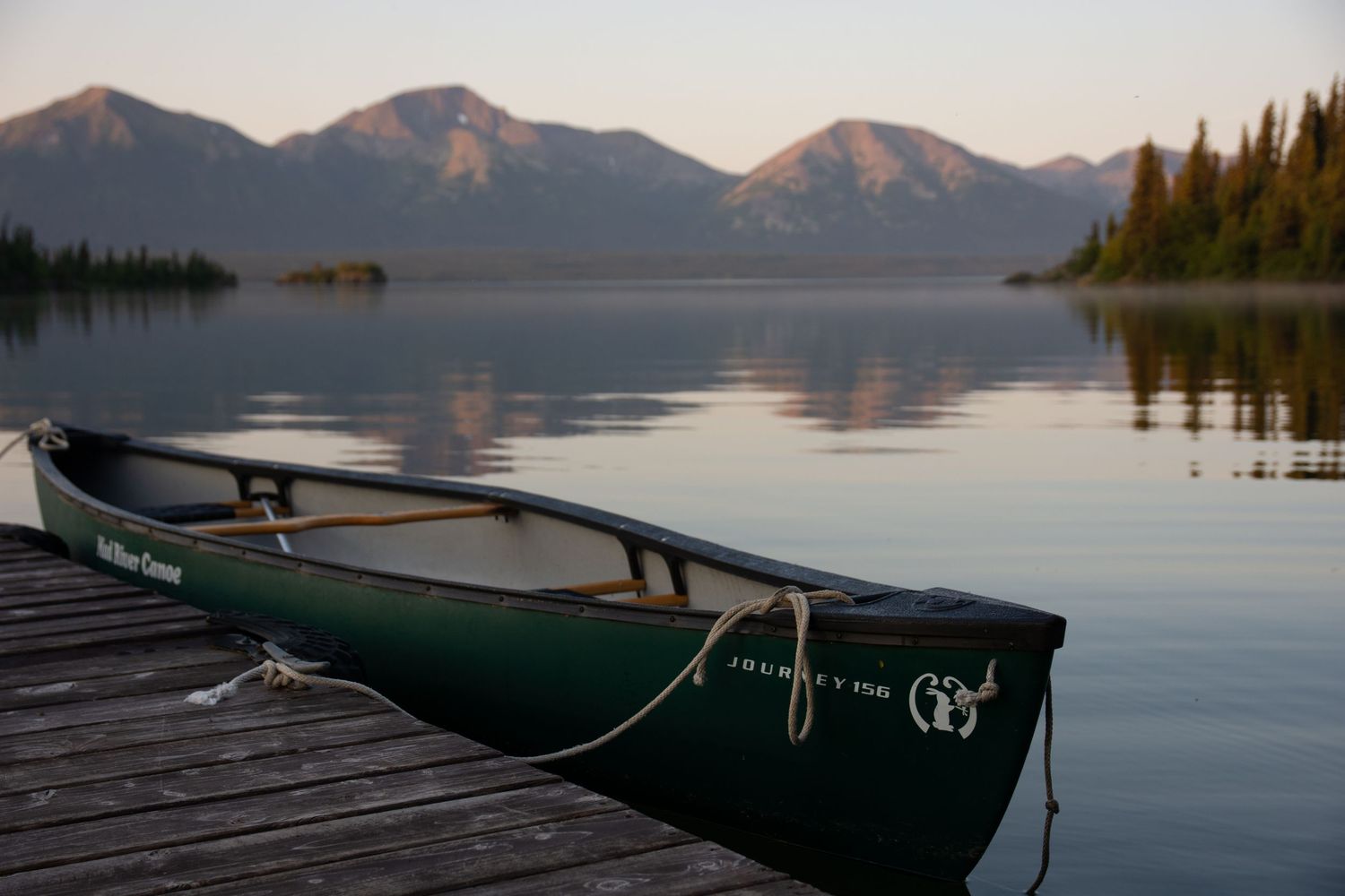 Green canoe tied to wooden dock on calm mountain lake at sunset.