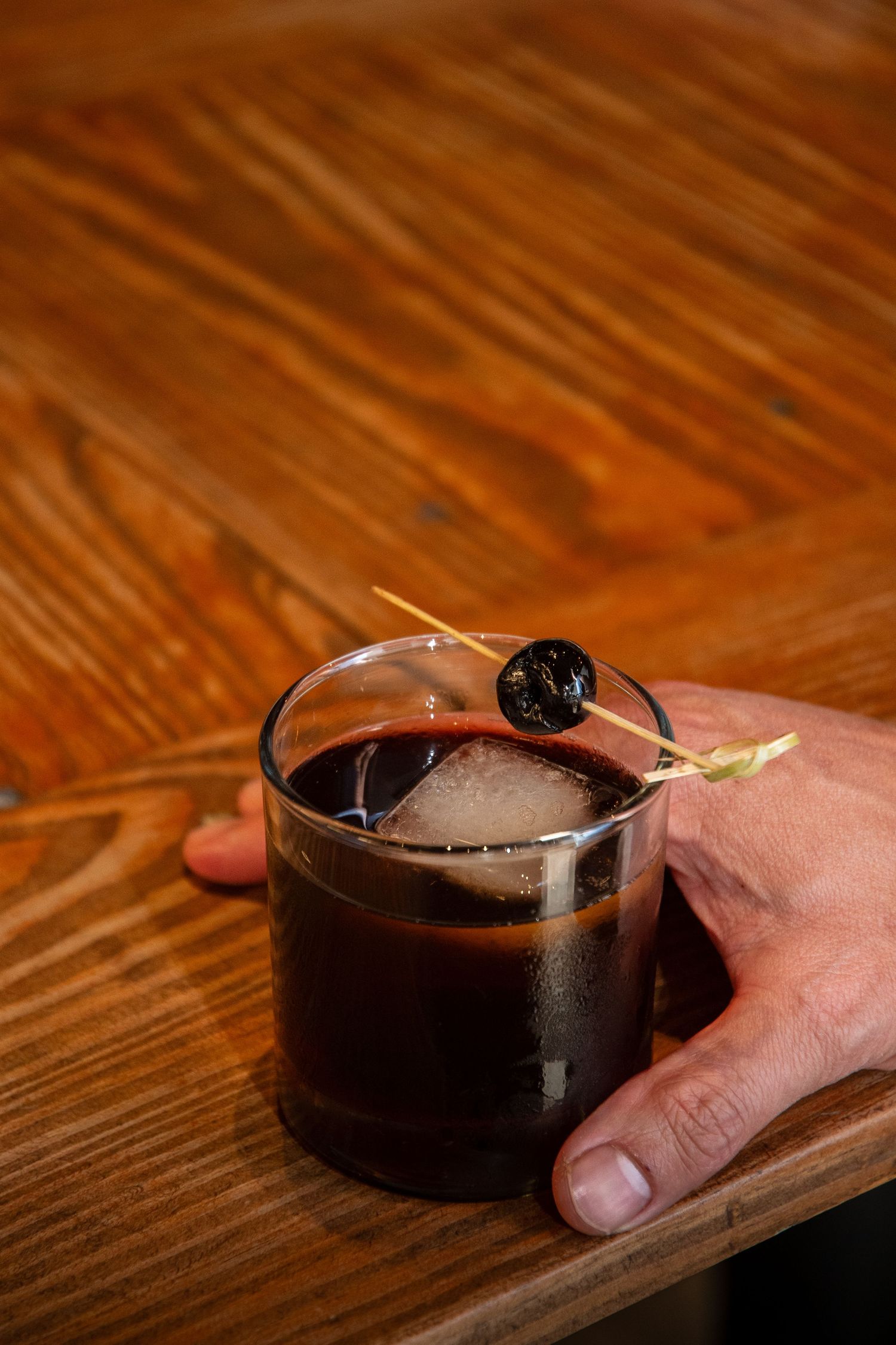 Dark cocktail with ice garnished with a cherry served on a wooden board.