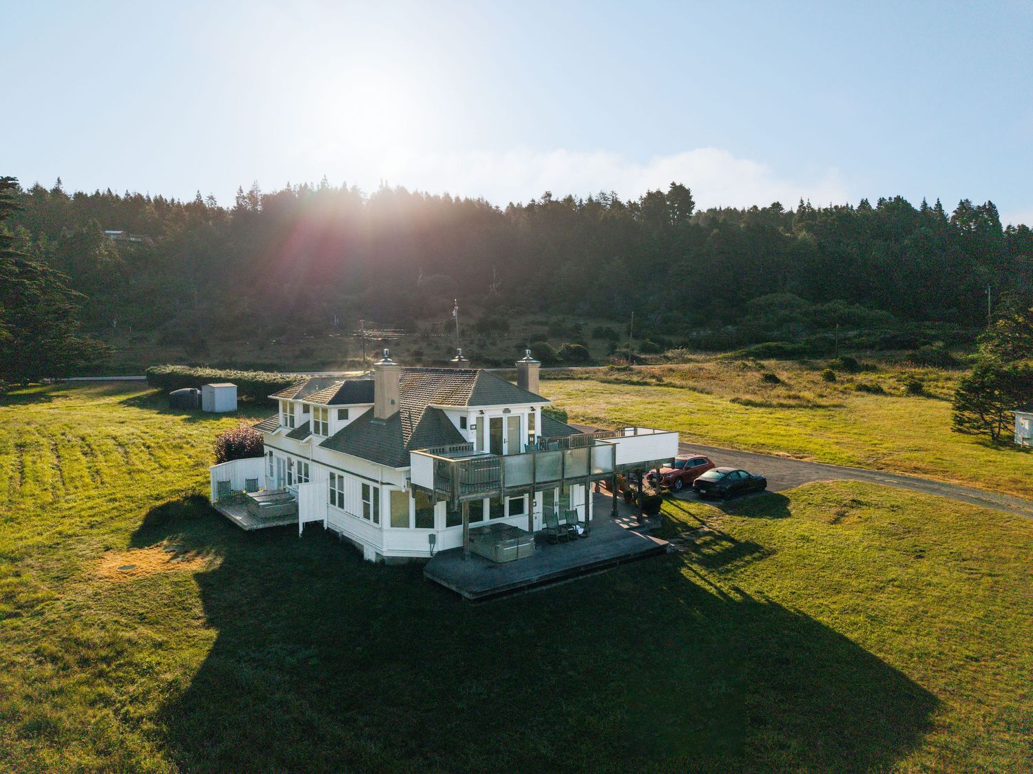 Aerial view of a white farmhouse surrounded by lush green fields with a dense forest and golden sunlight in the background.