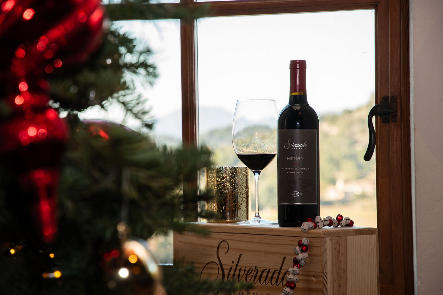 Wine bottles and glasses silhouetted against window with red holiday lights.