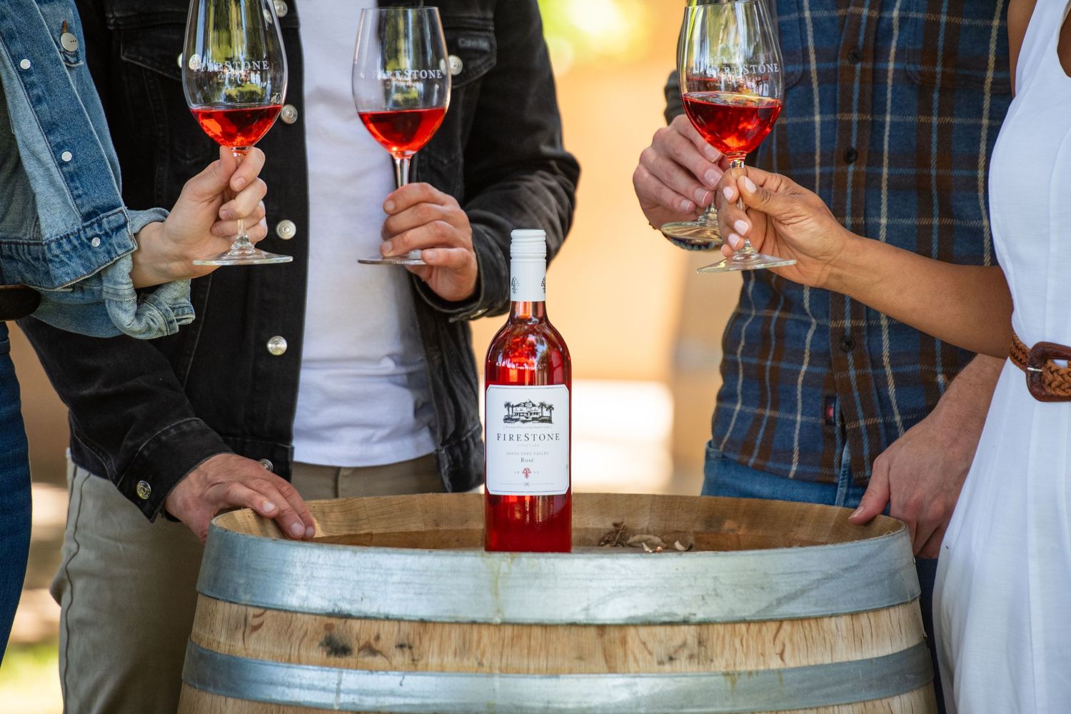 Three people toasting with rosé wine glasses over a wooden barrel with a bottle of rosé at a winery.