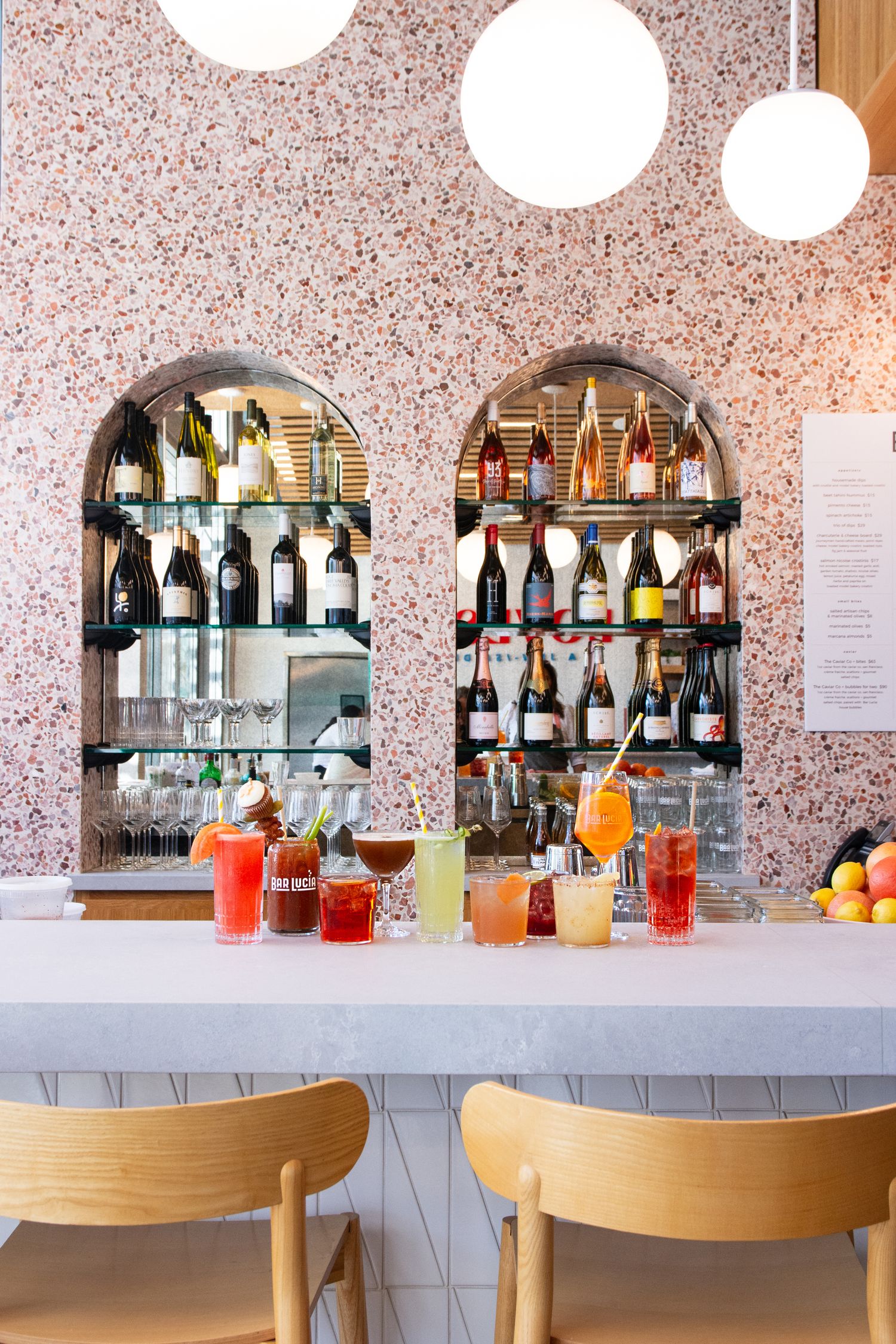 Bar counter with arched shelving displaying liquor bottles against pink terrazzo wallpaper.
