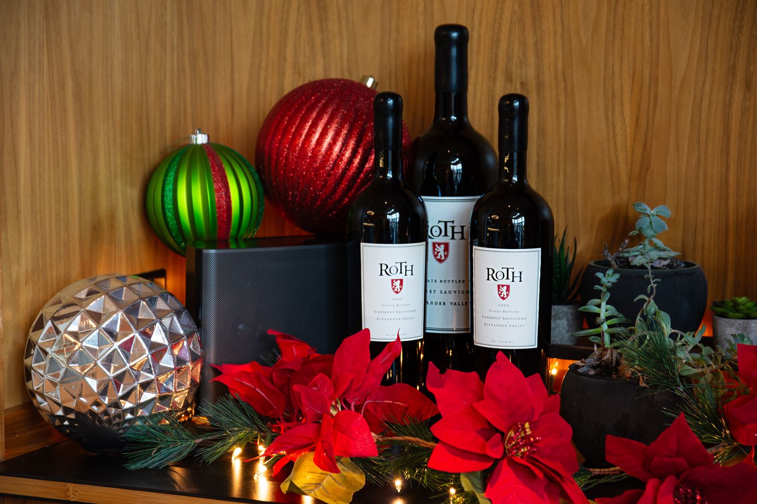 Festive holiday display with wine bottles, ornaments, and poinsettias arranged on a wooden surface.