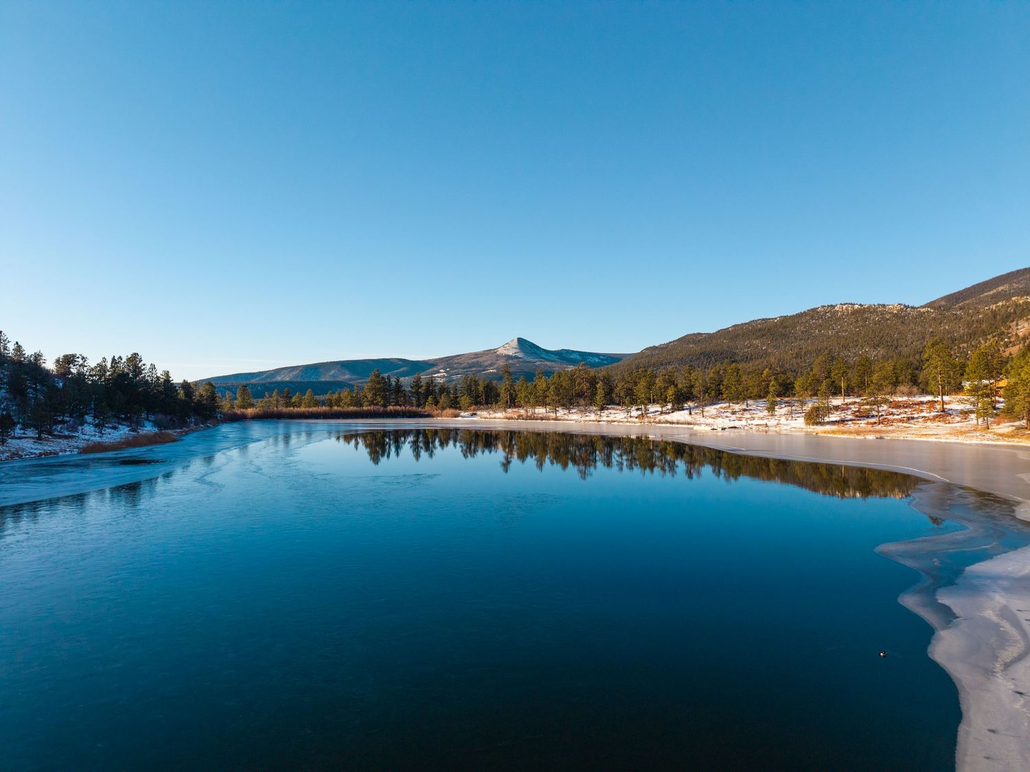Serene mountain lake with crystal-clear reflections, snow-dusted shores, pine trees, and a peak under a vivid blue sky.