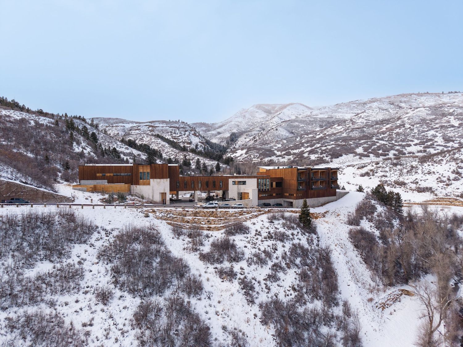 Snow-covered mountain landscape with modern luxury home nestled among winter hills and bare shrubs under overcast sky.