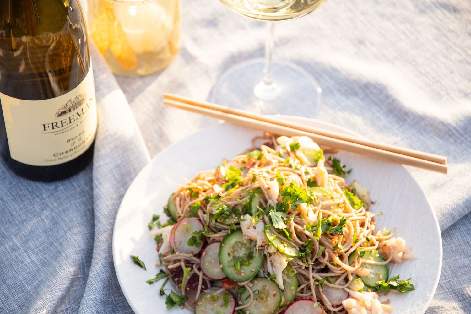 Asian noodle salad with fresh herbs served alongside a bottle of white wine on blue linen.