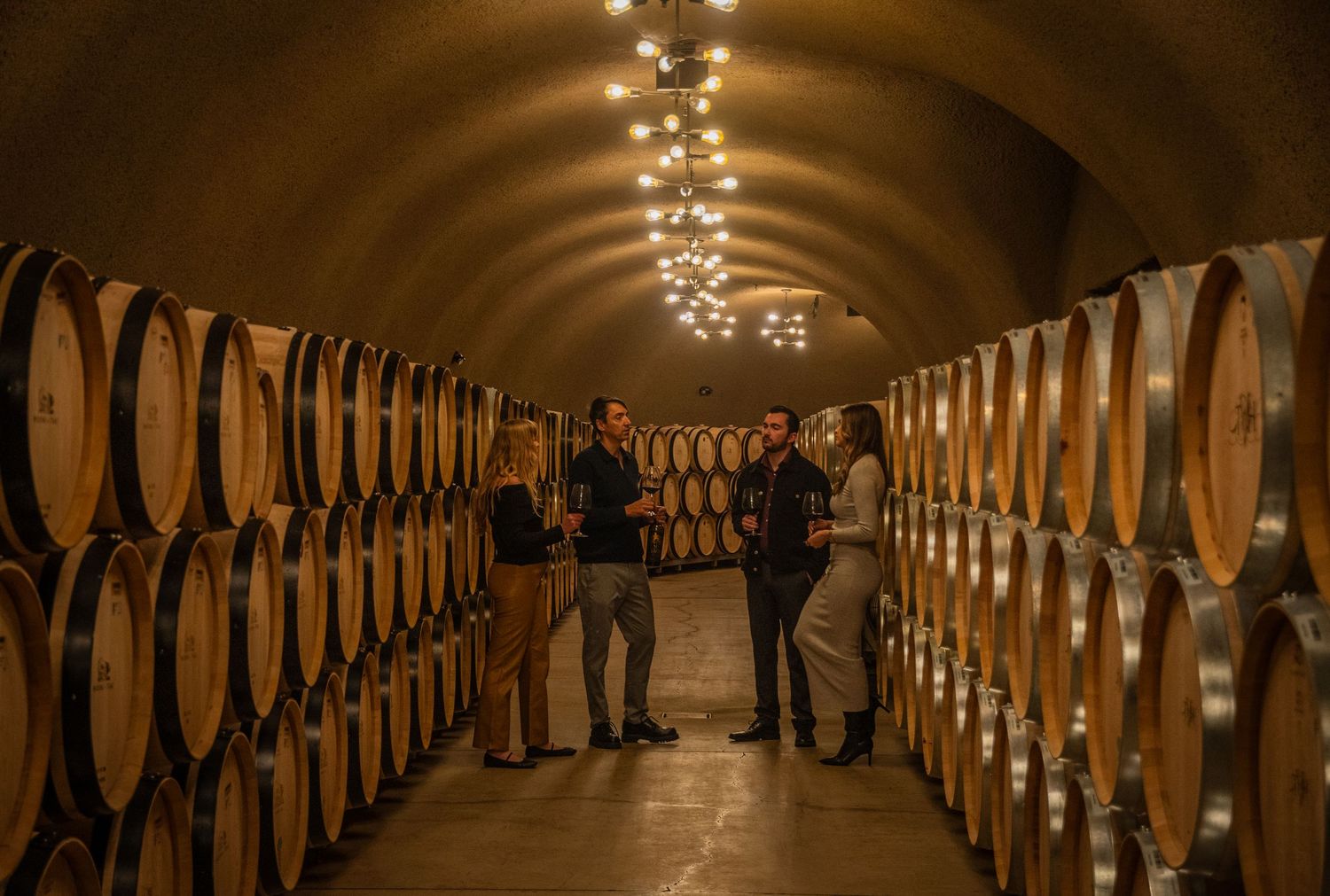 Long wine barrel cellar with dramatic lighting and arched stone ceiling.