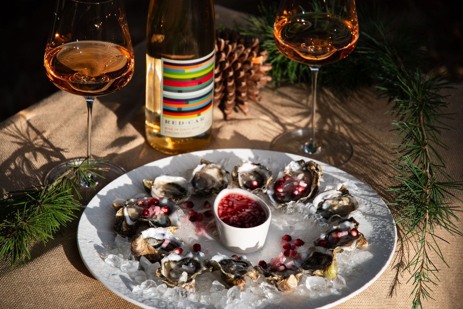 Fresh oysters on ice with pomegranate sauce and wine glasses arranged on a rustic table with pine decorations.