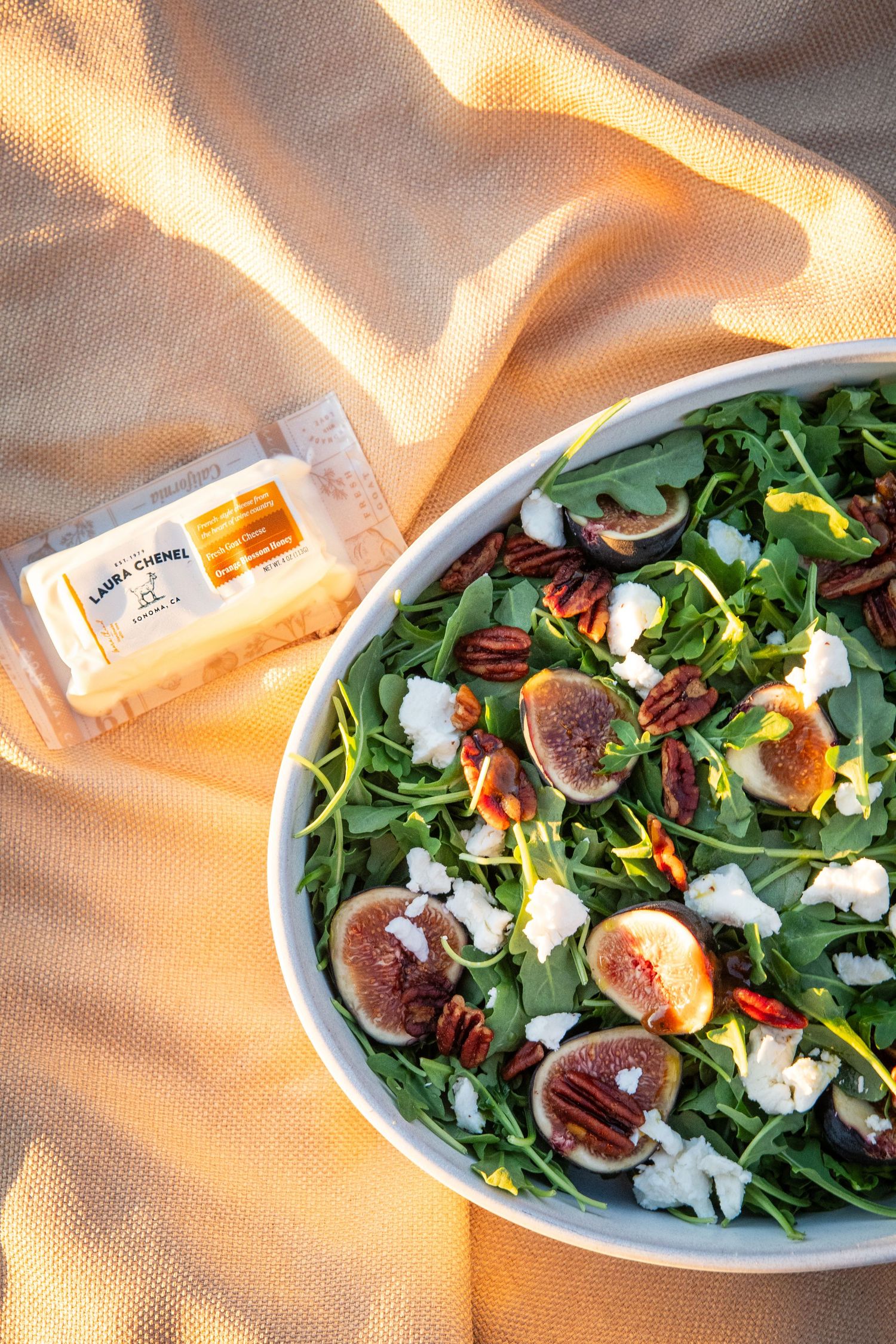 Fresh fig and goat cheese salad with mixed greens in white bowl on sunlit fabric.