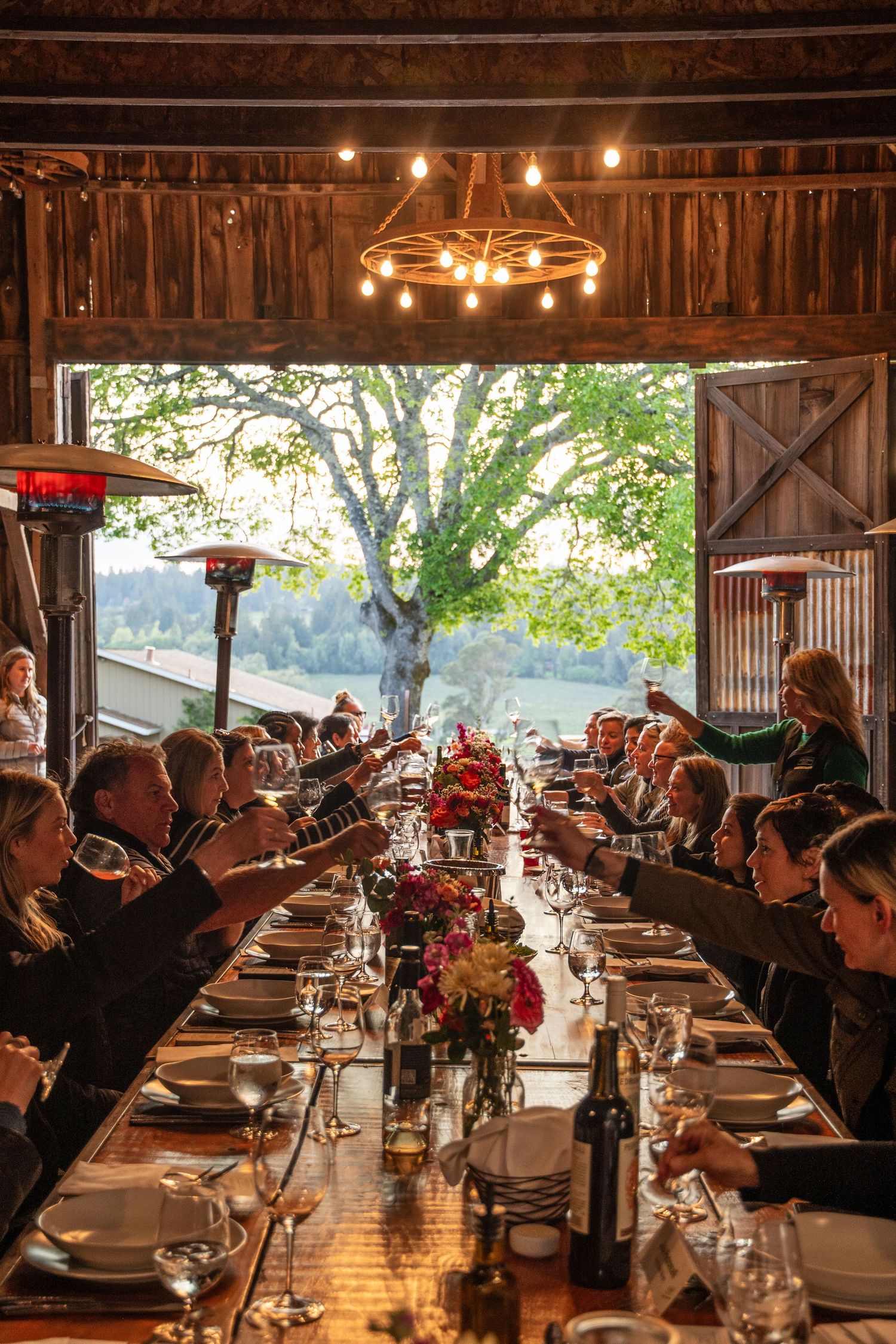 Diners raise their glasses in a toast at long tables inside a wooden barn-style venue with natural lighting.