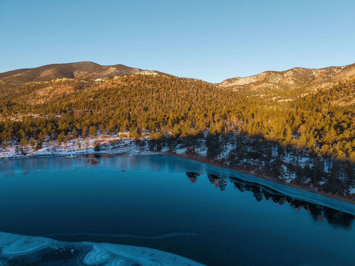 Aerial view of a serene partially frozen lake surrounded by pine forests and mountains at golden hour in winter.