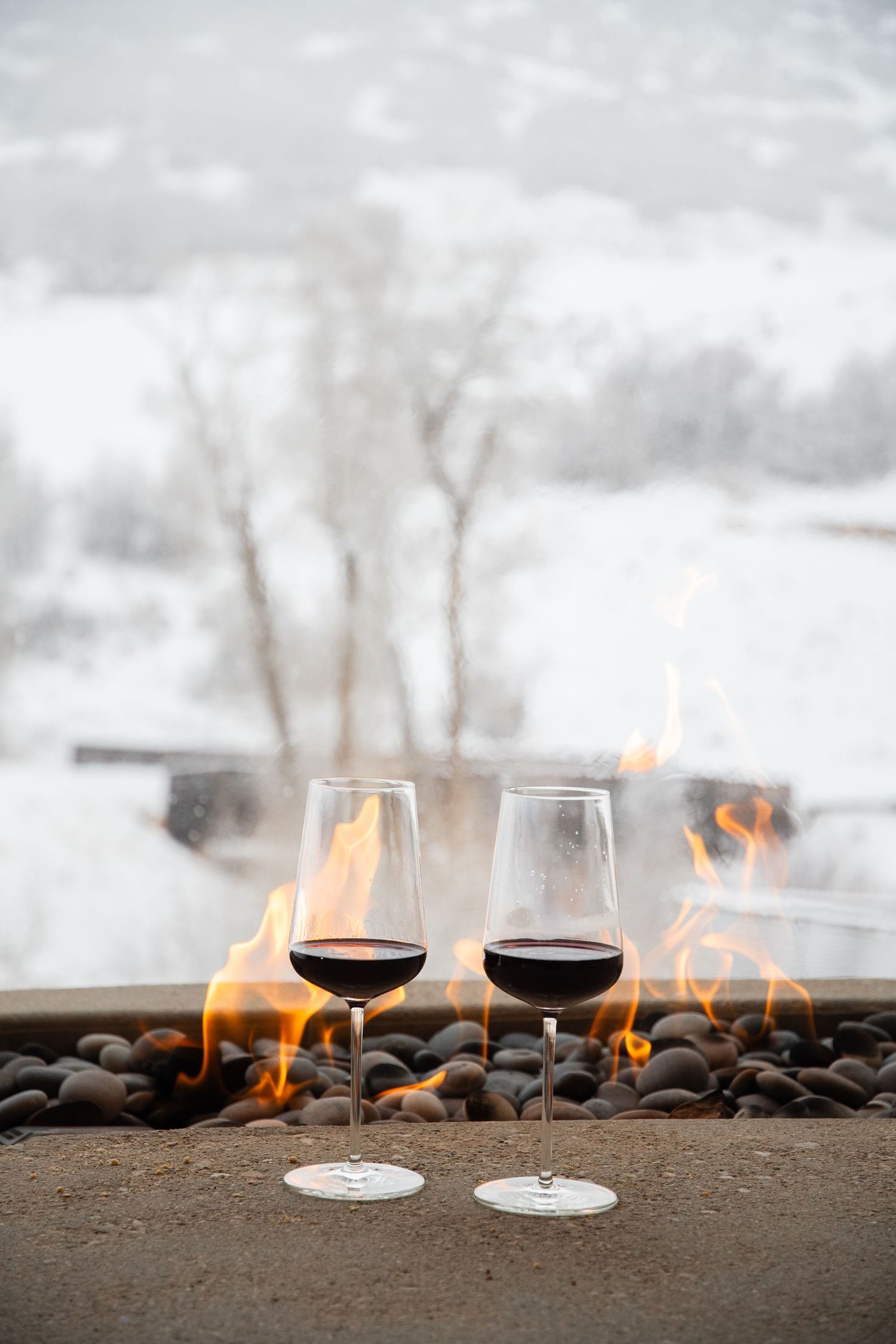 Two wine glasses set against a snowy backdrop with a fire feature burning behind them.