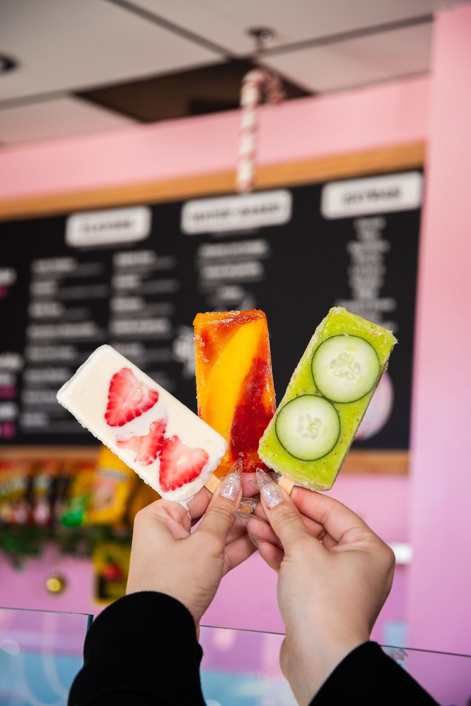 Colorful popsicles with fruit and cucumber slices held up against a pink wall.