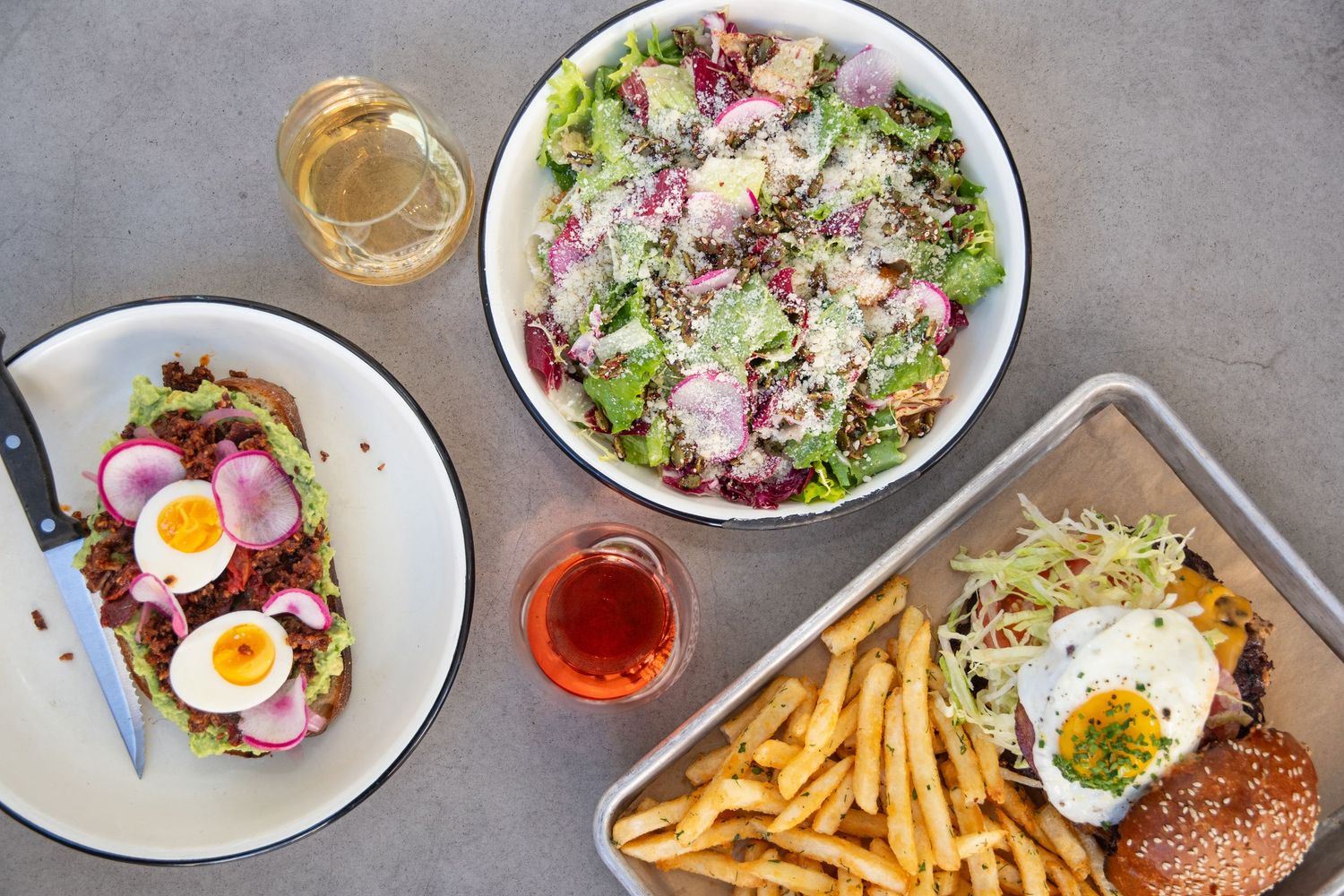 Overhead view of restaurant dishes including salads, burgers, and french fries arranged on a gray surface.